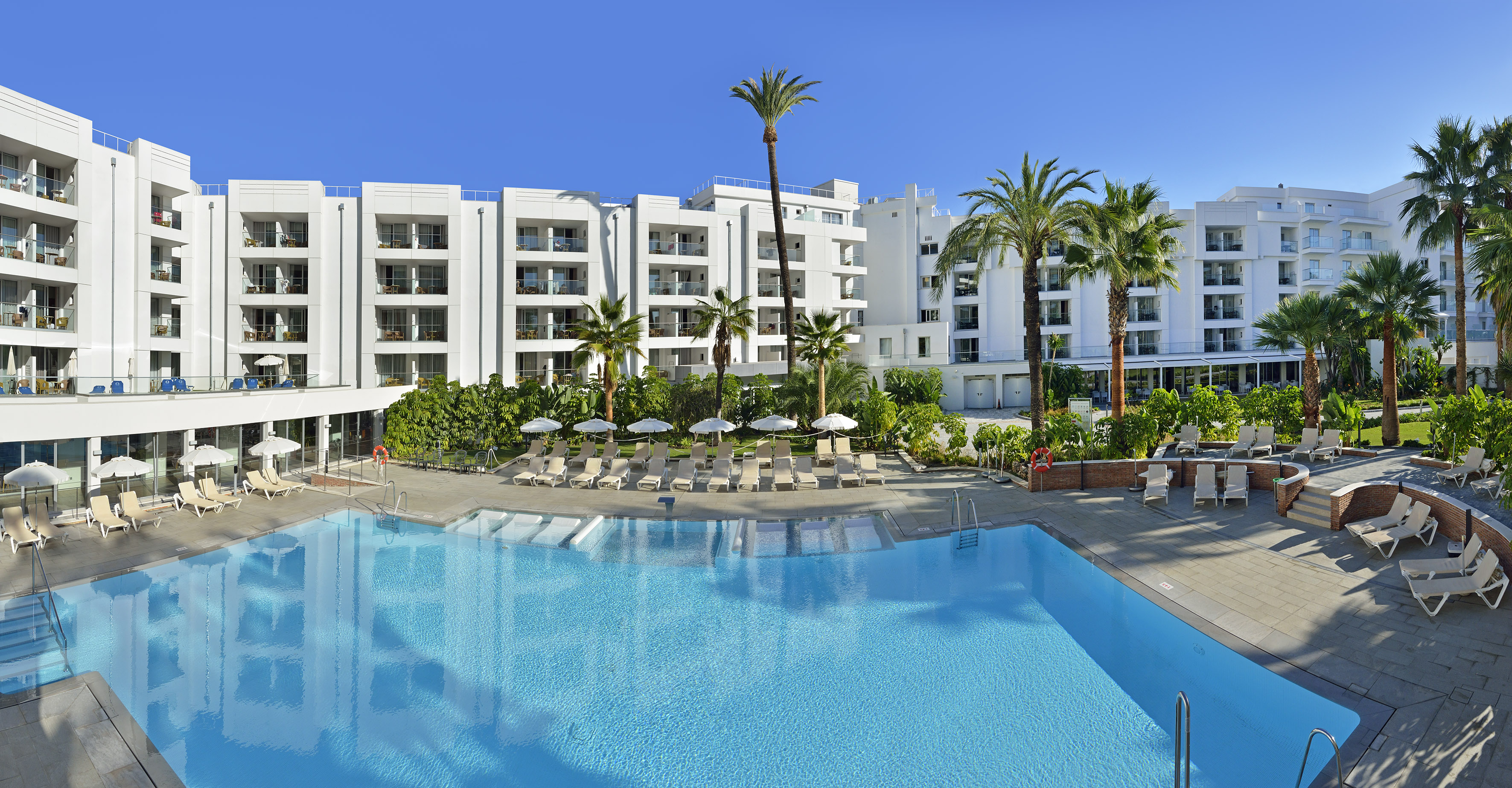 White resort hotel, large swimming pool, palm trees, and sun loungers.