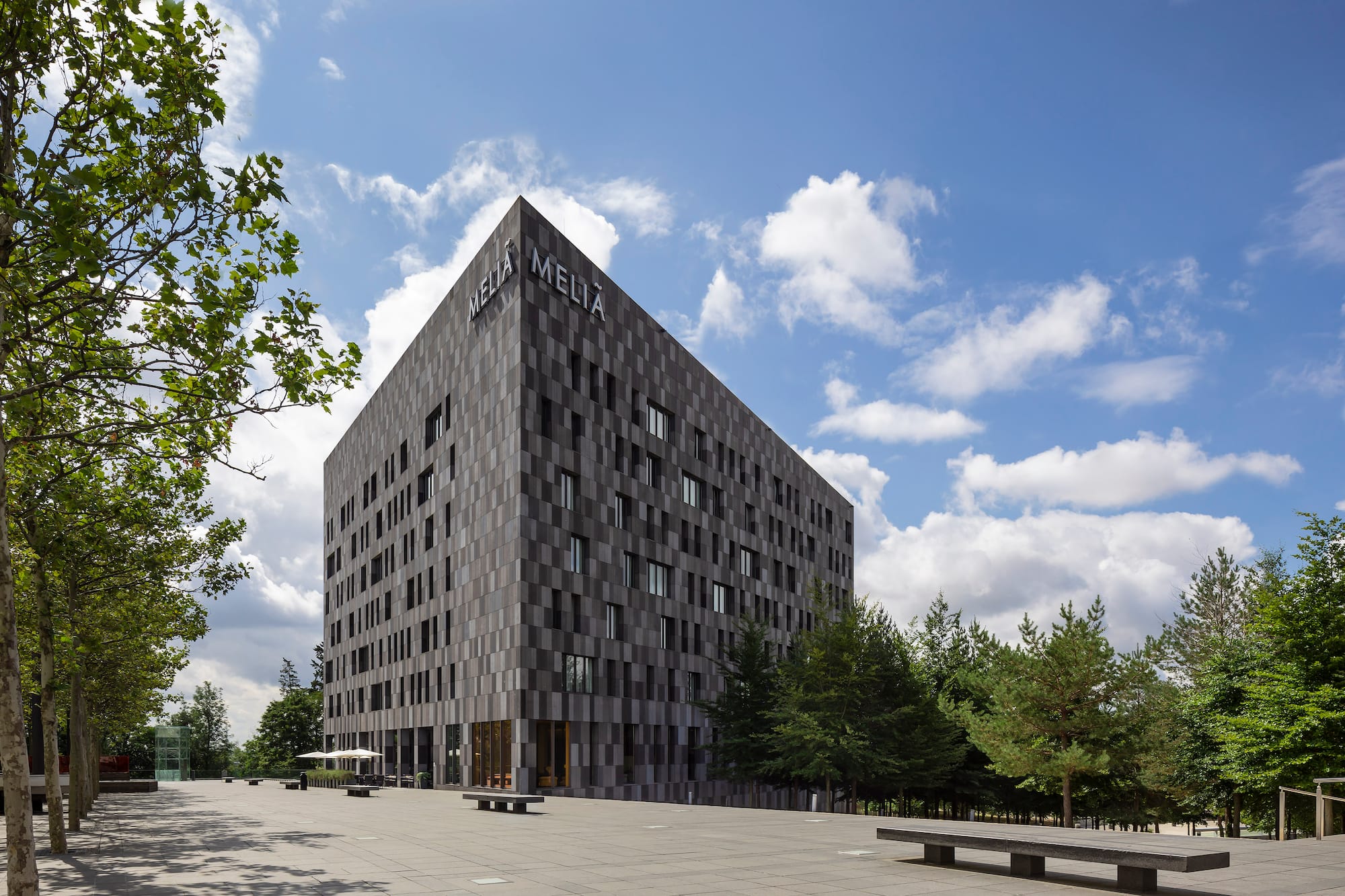 Meliá hotel, modern dark gray building, paved plaza, green trees, blue sky.
