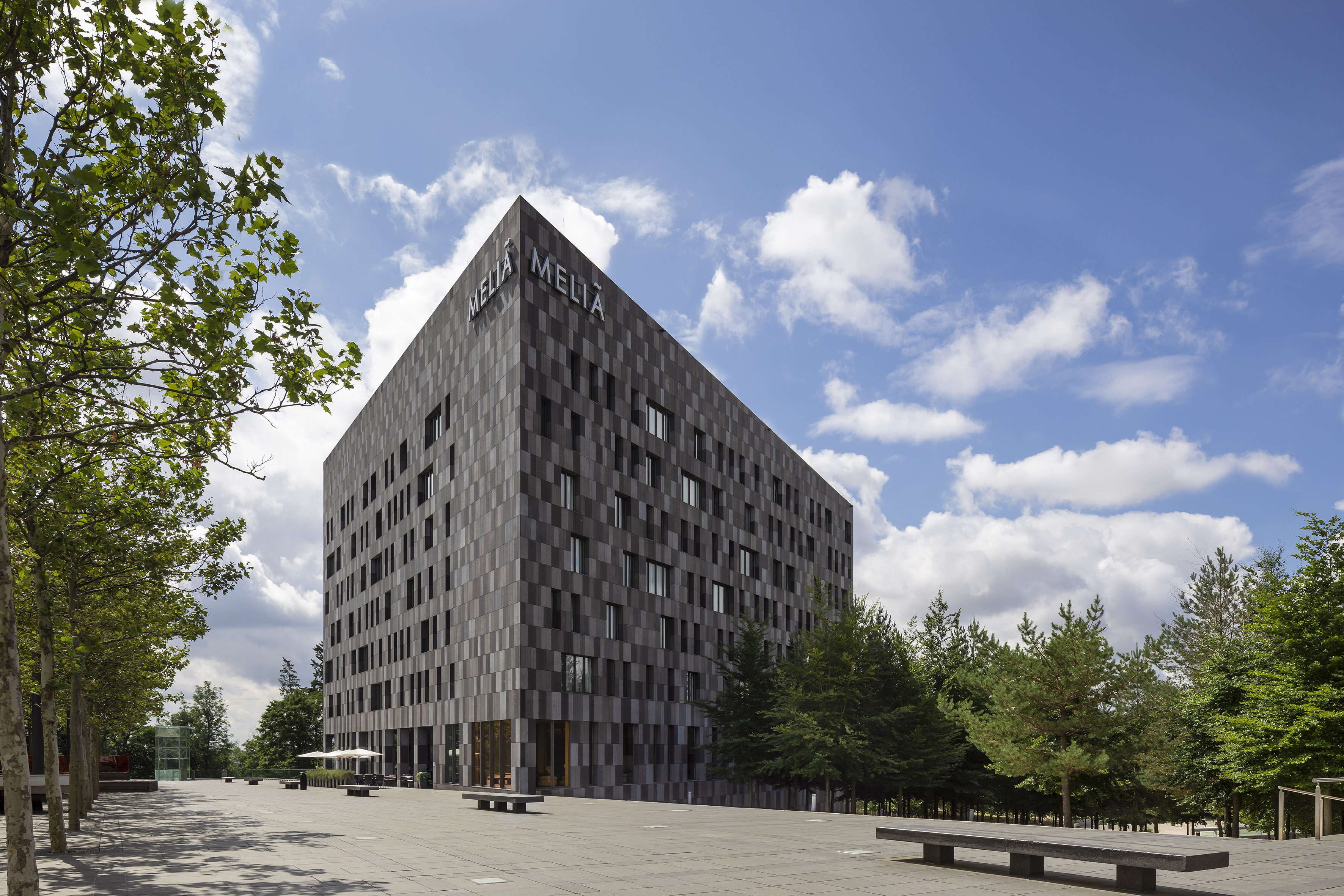 Meliá hotel, modern dark gray building, paved plaza, green trees, blue sky.