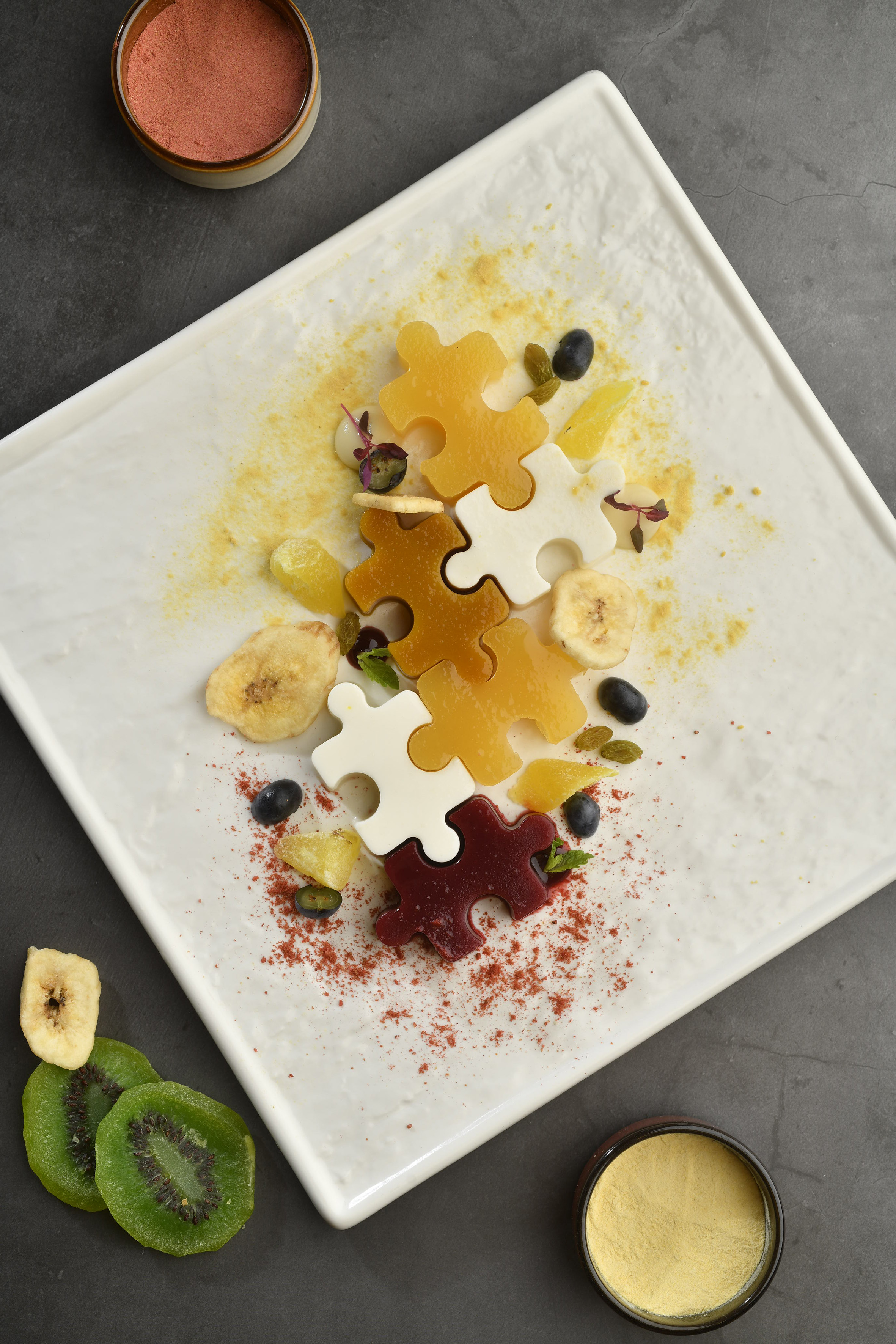 Puzzle-shaped fruit jellies with dried fruit, berries, and spice powders on white plate.