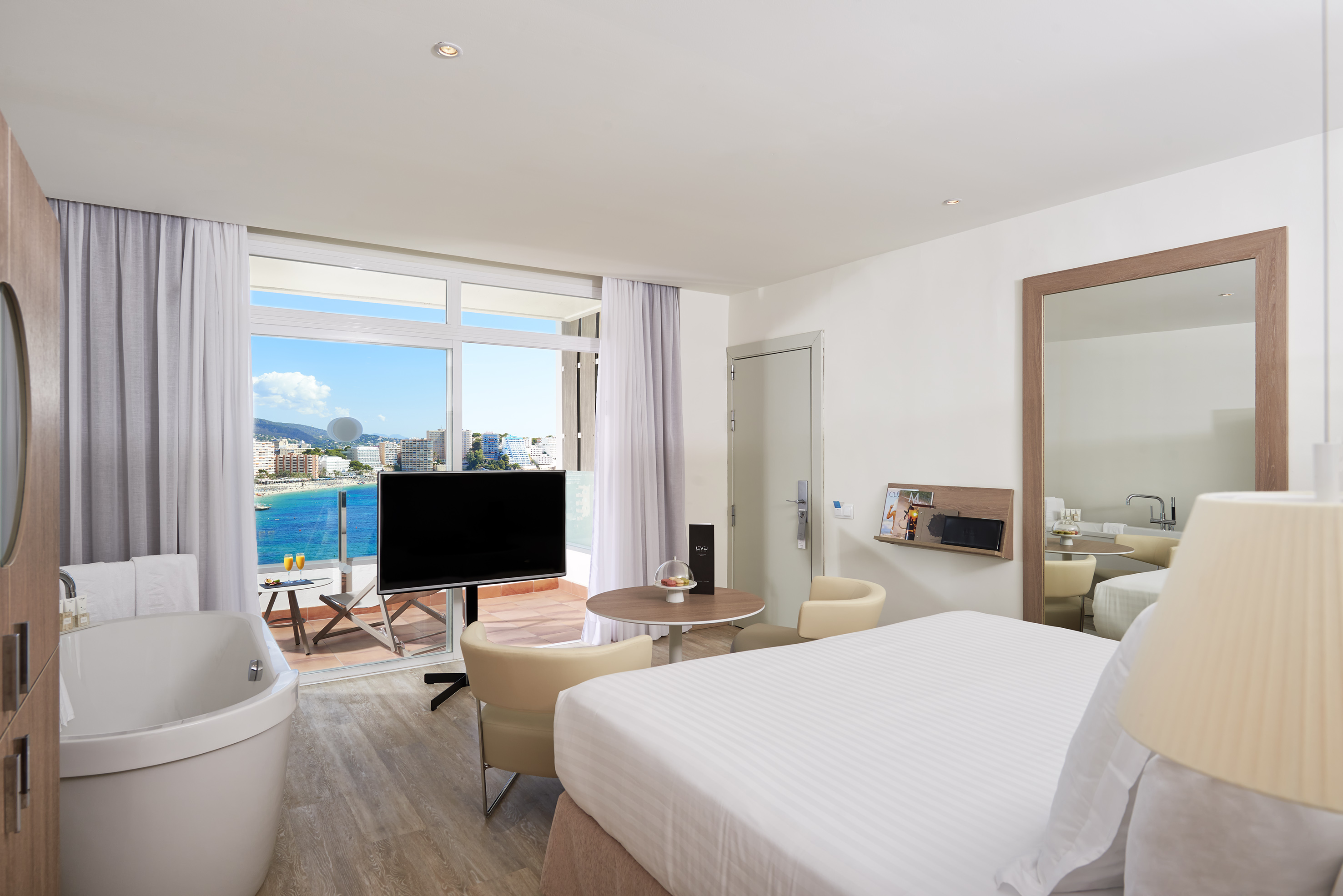 Modern hotel room with sea view balcony, large bed, TV, and corner bathtub.