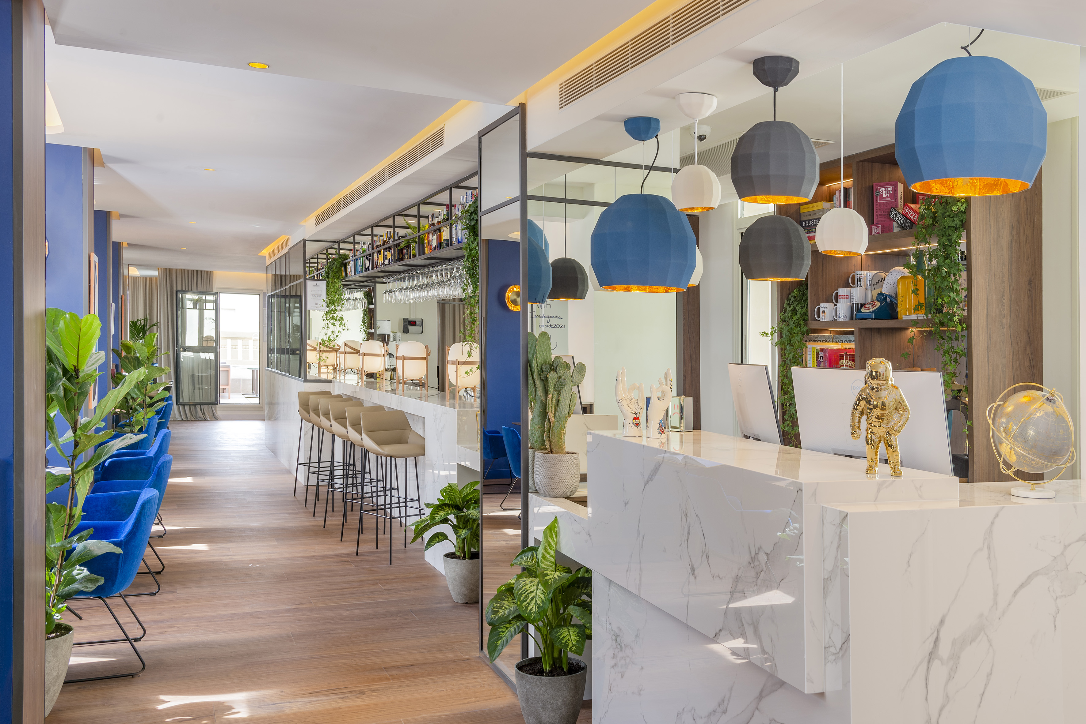 Modern hotel lobby bar with marble counter, blue chairs, plants, and geometric pendant lights.
