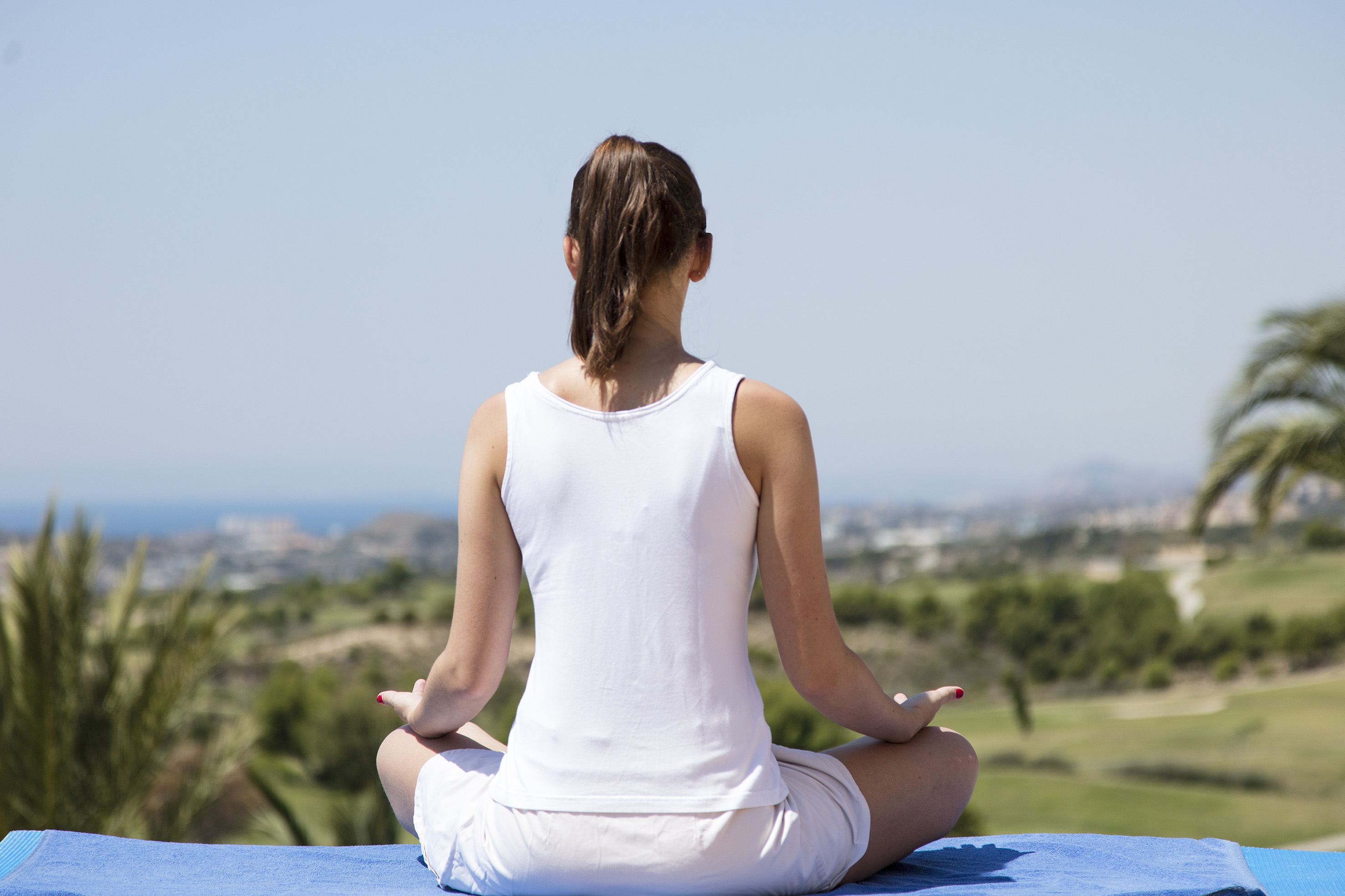 Woman in white meditating outdoors on blue mat, scenic ocean & green landscape.