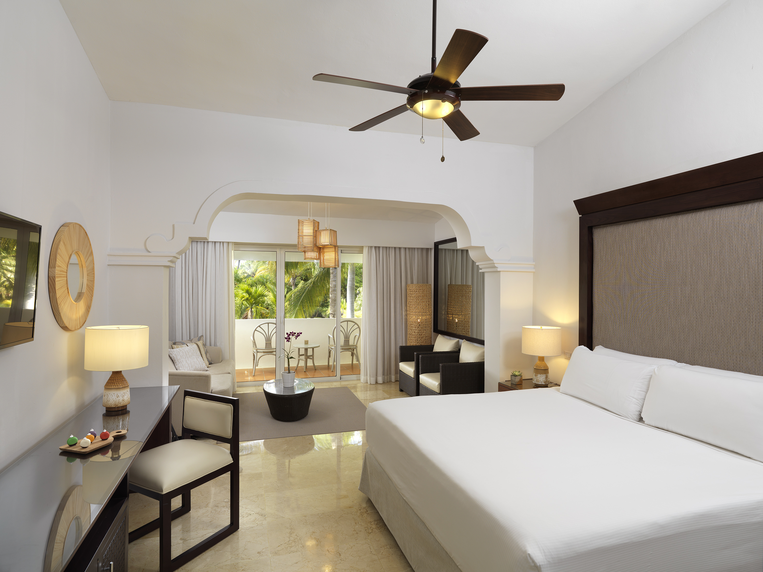 Resort suite with king bed, ceiling fan, desk, and balcony overlooking tropics.