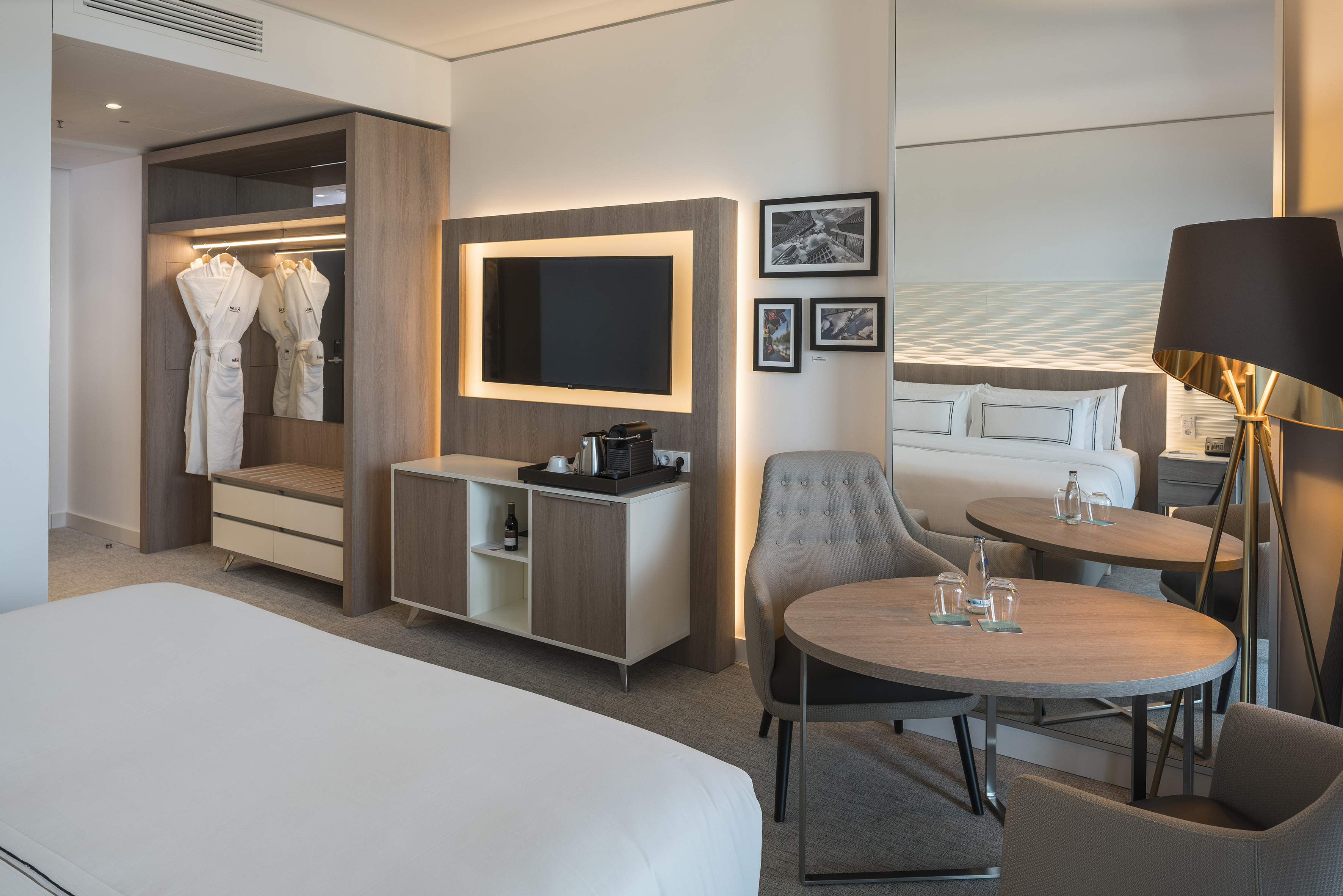 Modern hotel room interior with bed, wall TV, open wardrobe, and dining area.