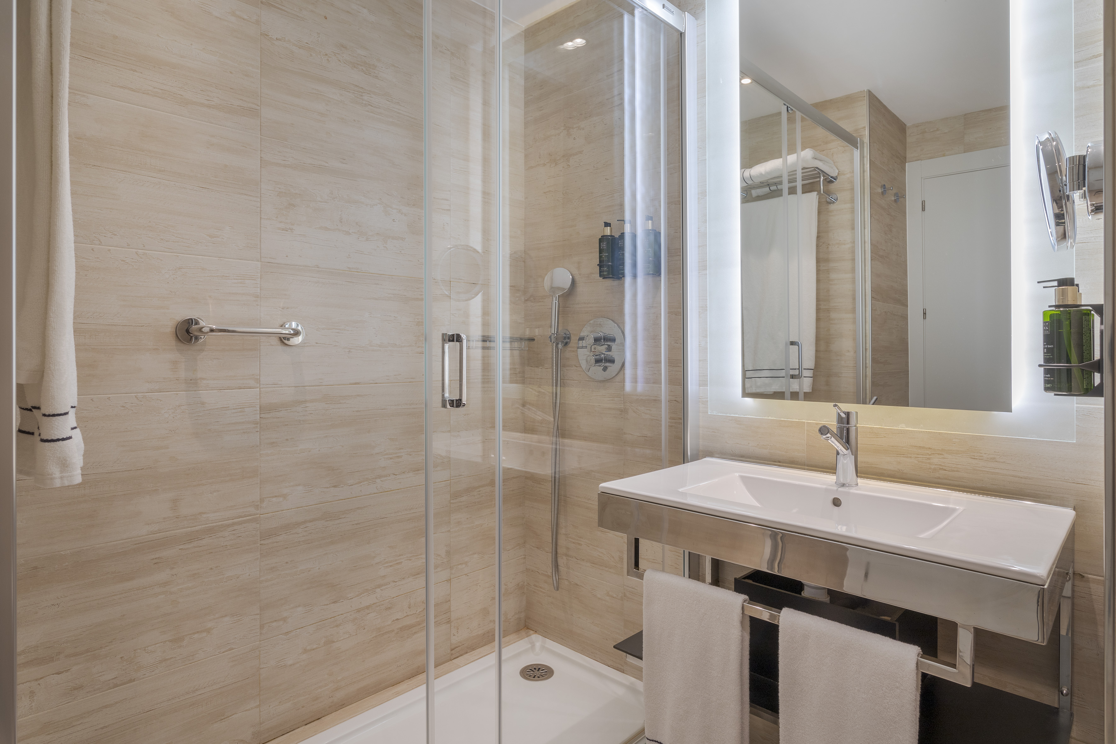 Modern hotel bathroom with glass shower, lighted mirror, and beige tiles.