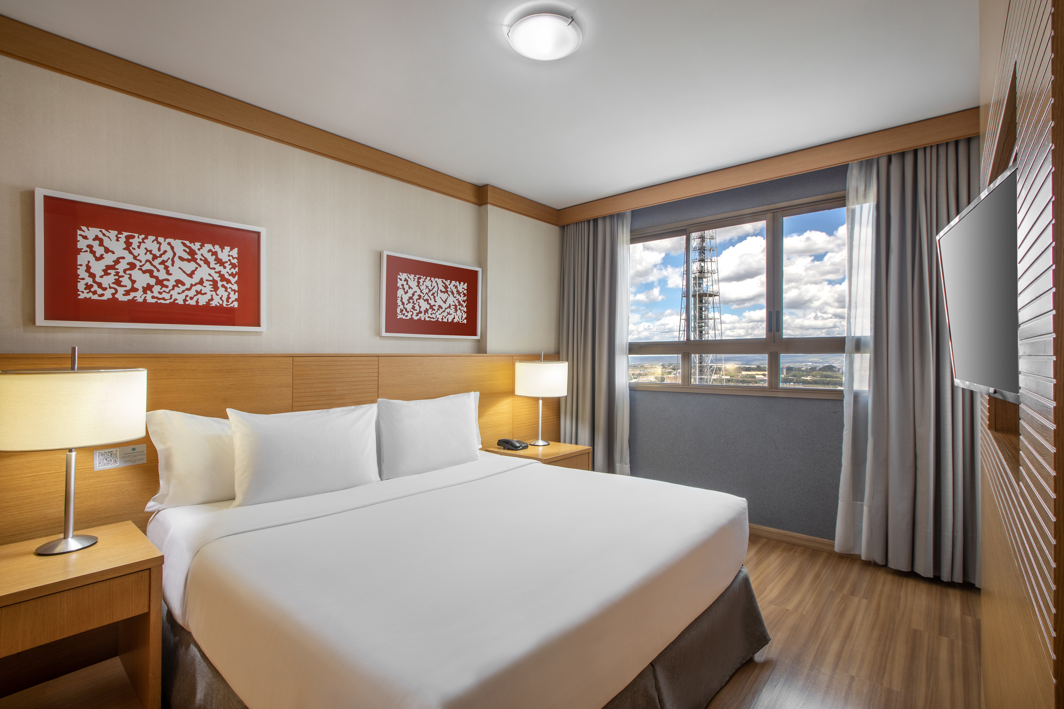 Modern hotel room with king bed, bedside lamps, city view window, and TV.