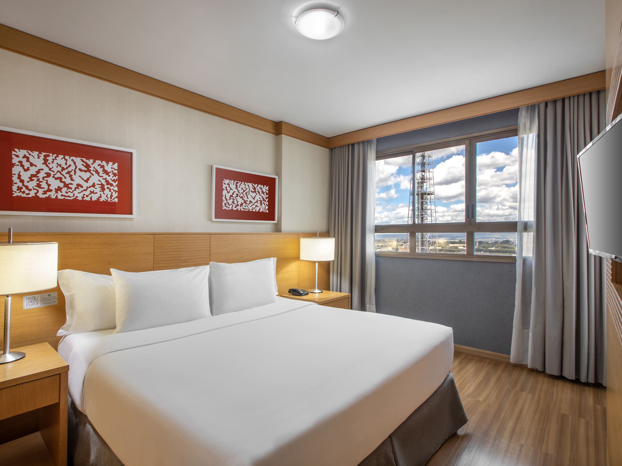 Modern hotel room with king bed, bedside lamps, city view window, and TV.