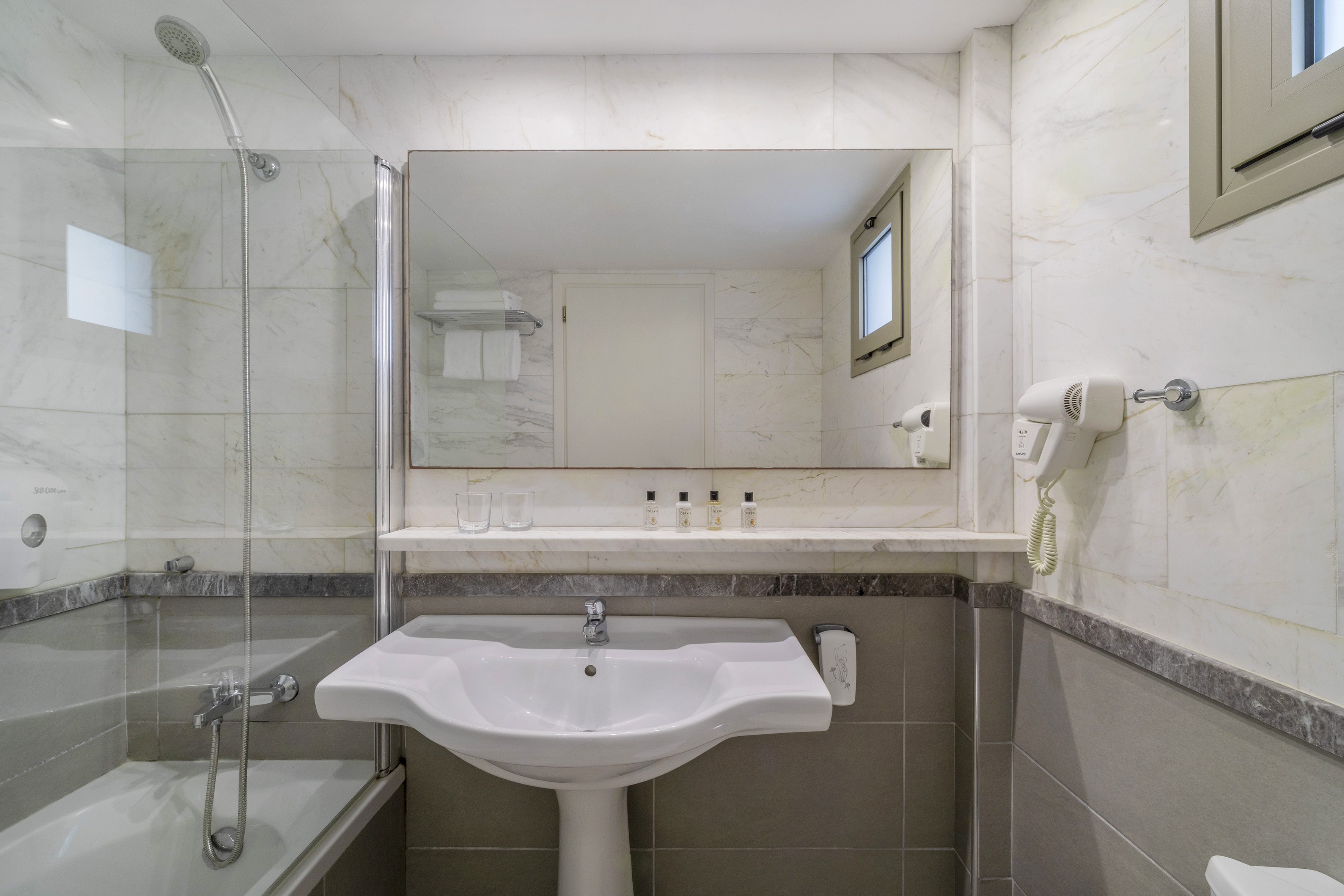 Hotel bathroom with white marble, sink, shower-tub, large mirror, and amenities.