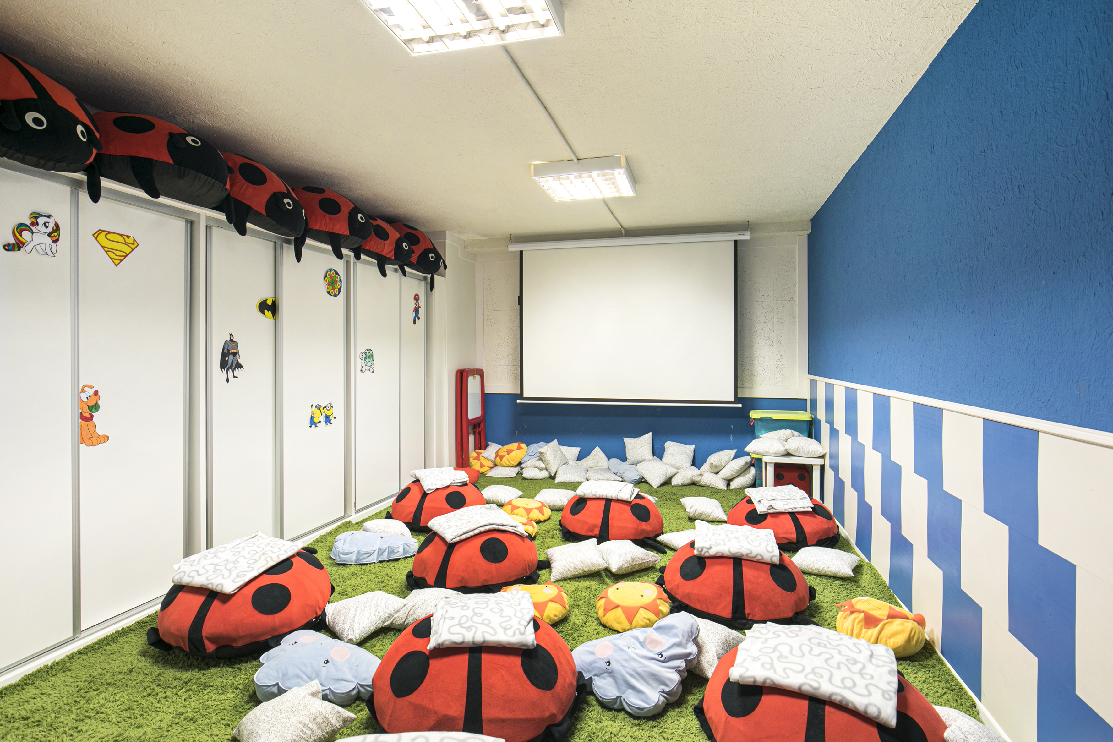 Kids' play area with red ladybug floor cushions on green carpet, projector screen.