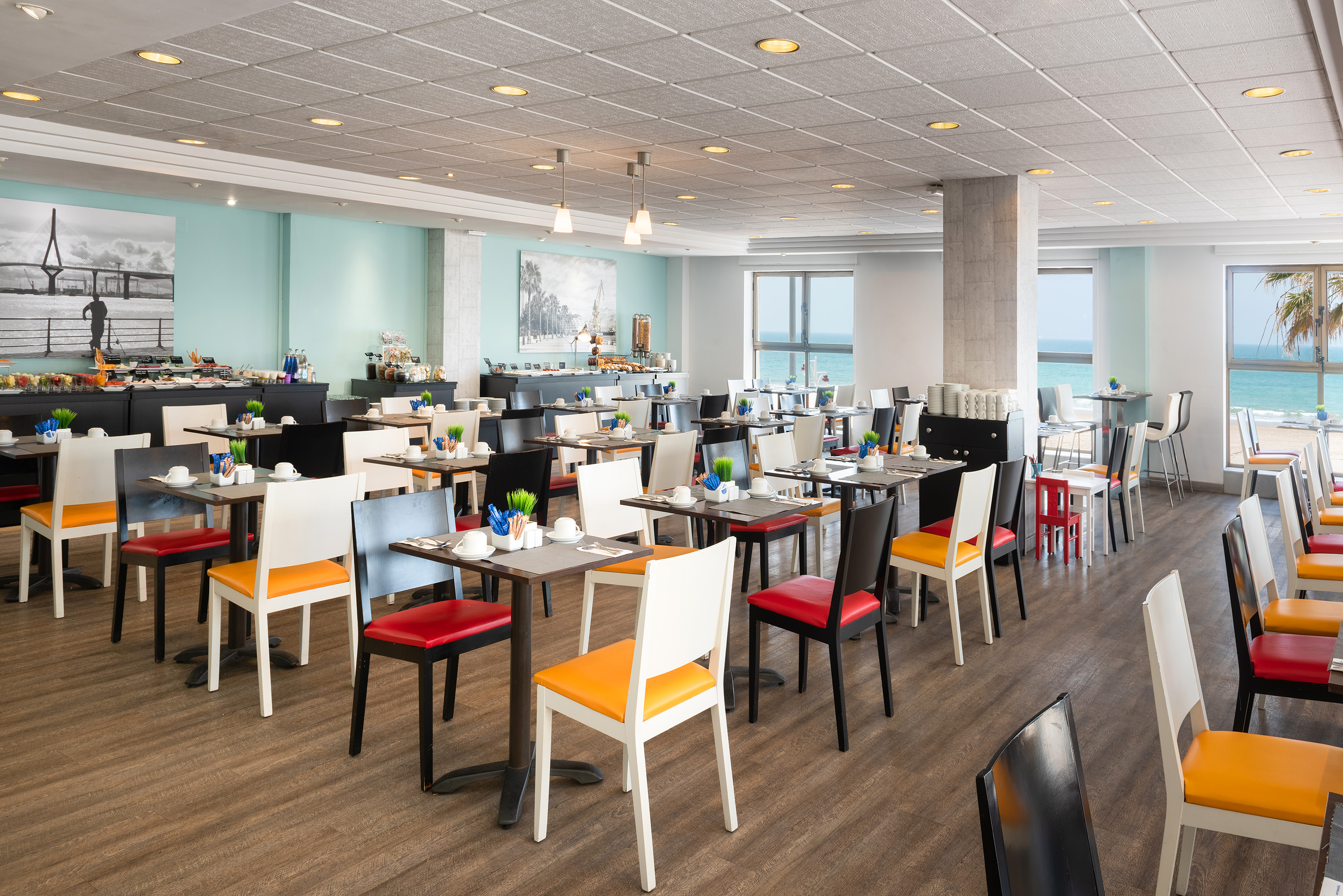 Modern hotel restaurant with beach view, colorful chairs, and buffet.
