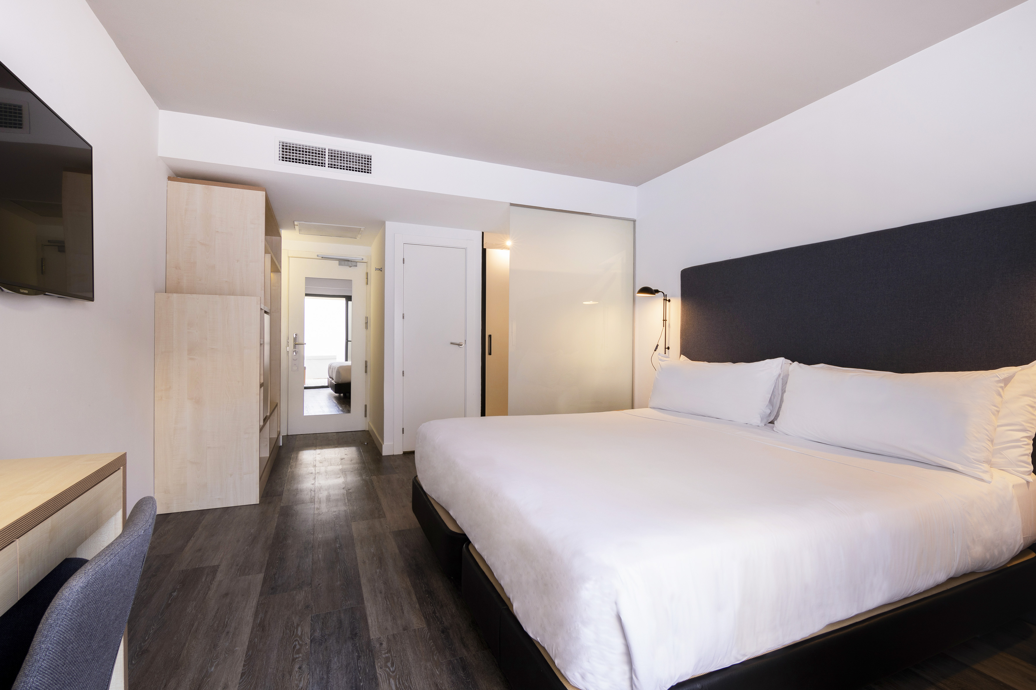 Modern hotel room with king bed, white linens, dark headboard, wood floor.