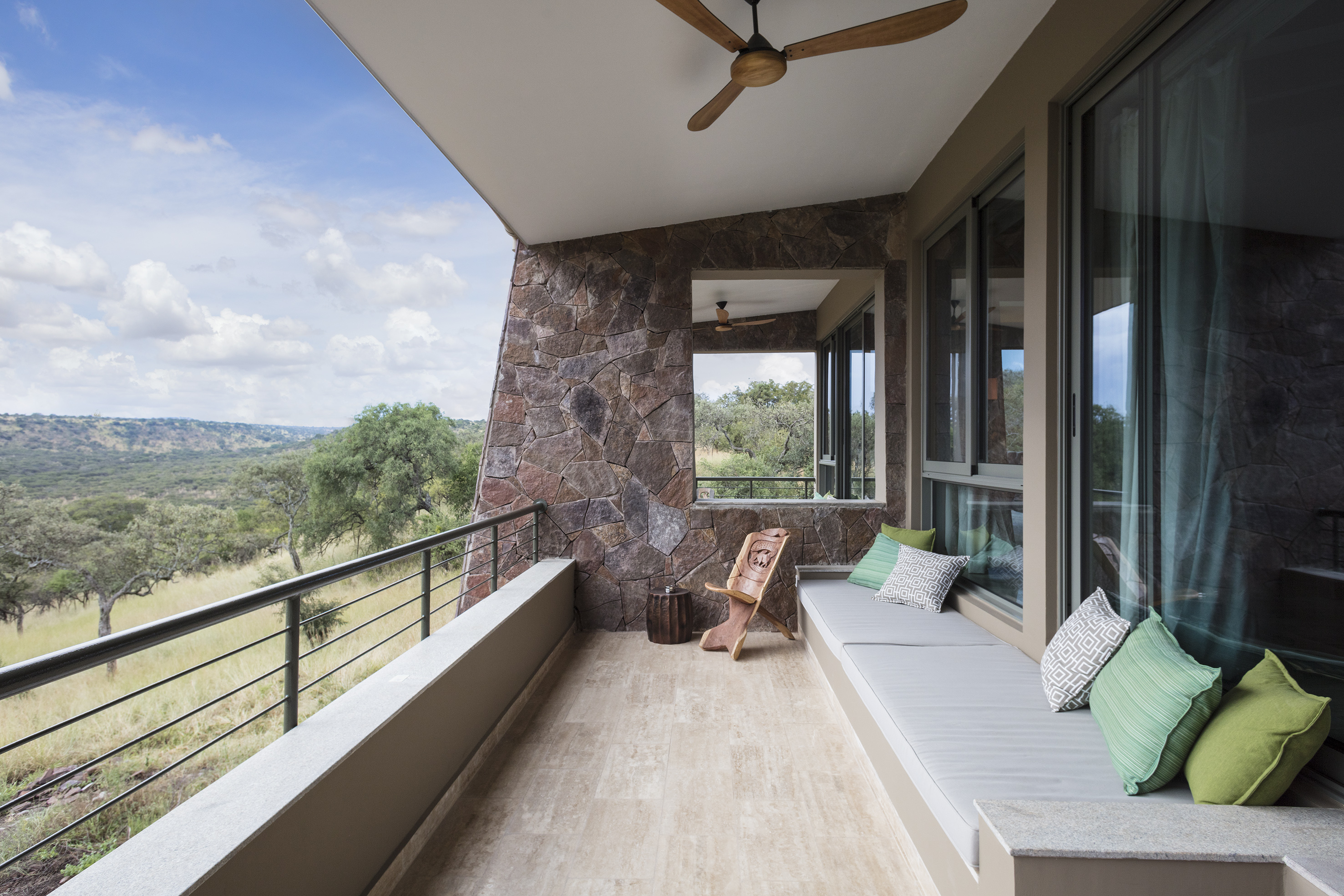 Lodge balcony with savanna view, stone wall, green cushions, and wooden ceiling fan.