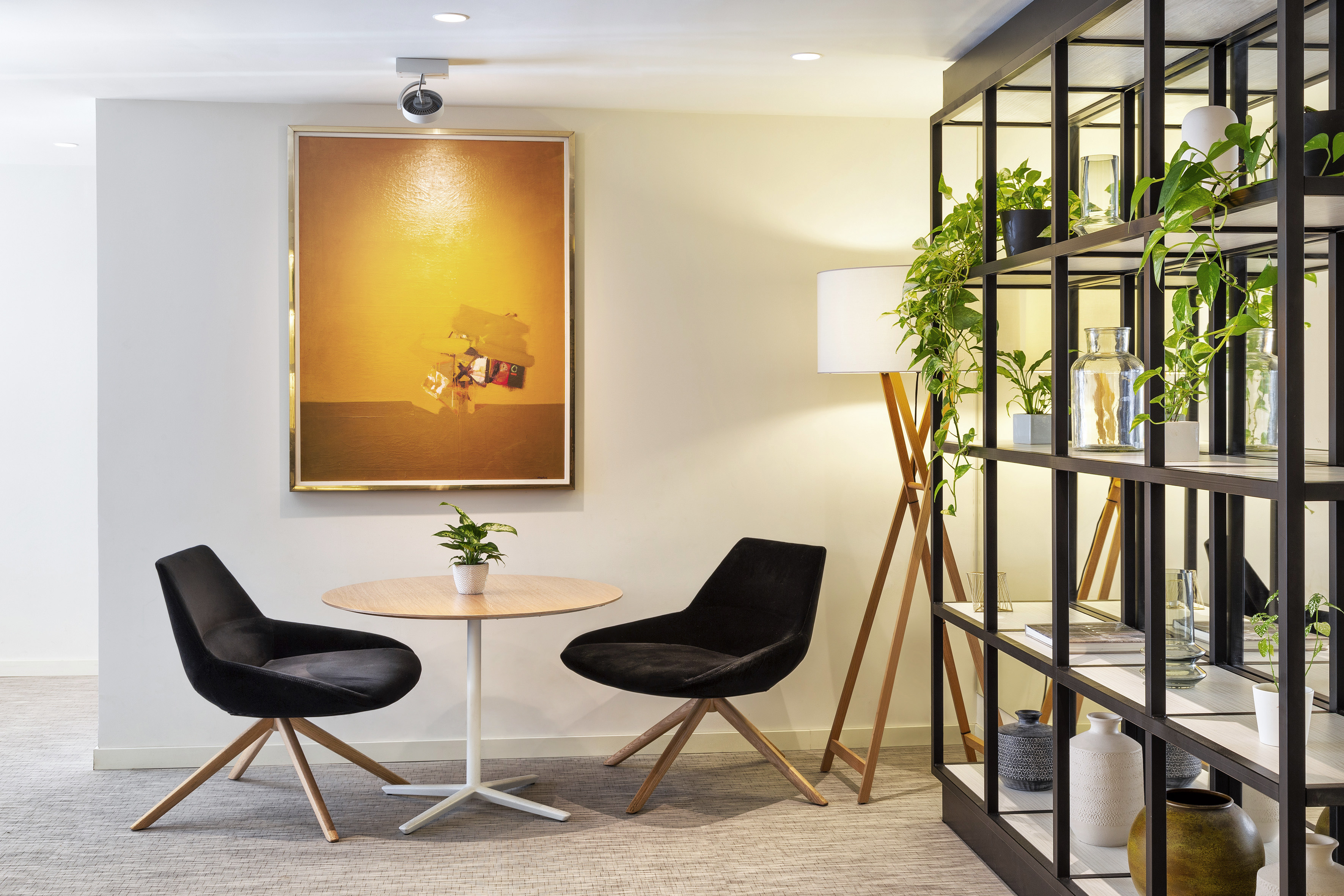 Modern lounge with black chairs, round table, abstract art, and plant-filled shelving.