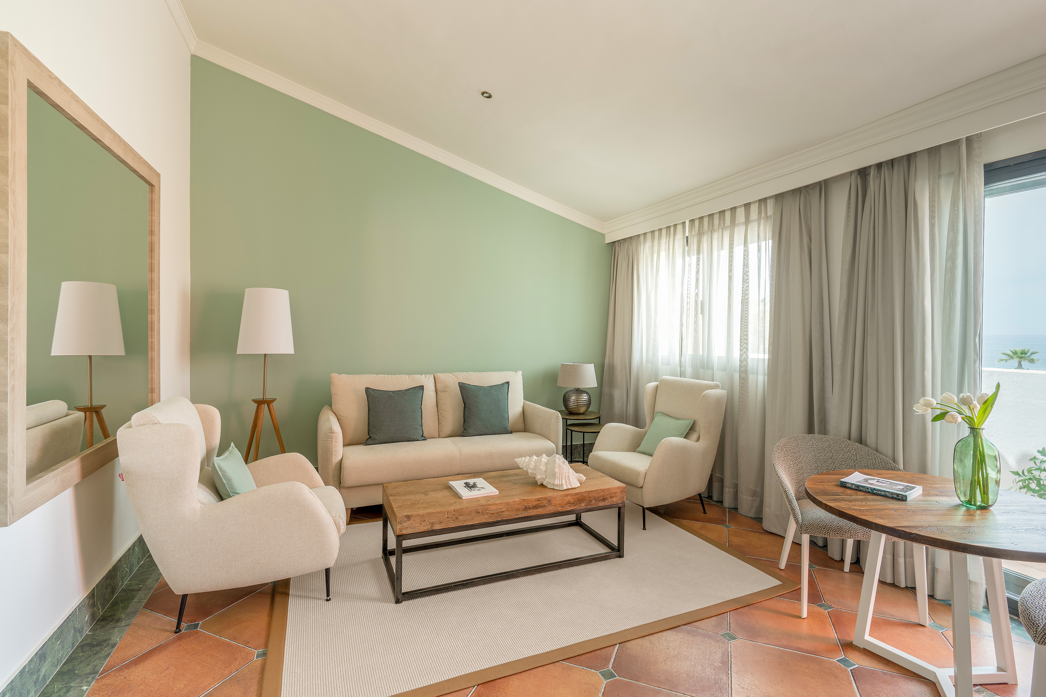 Cozy hotel suite lounge with green walls, cream sofa, wood table, ocean view.