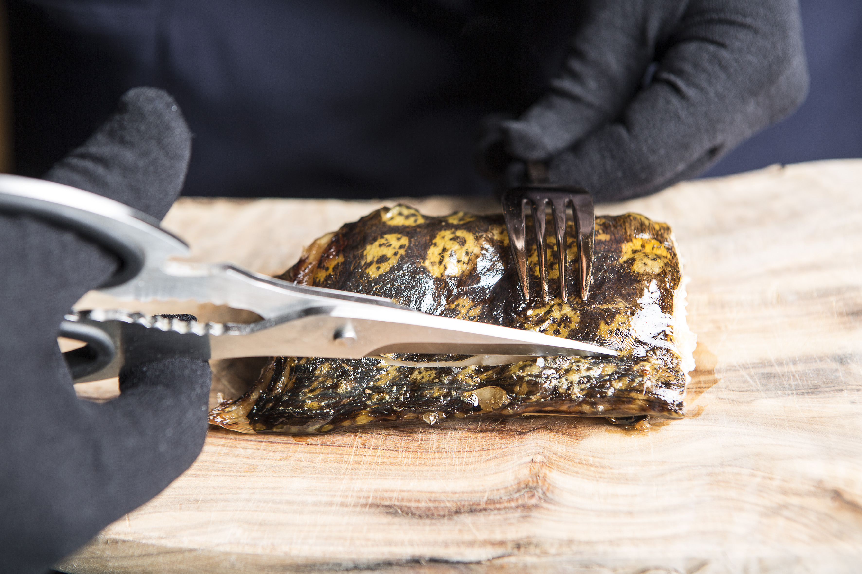 Gloved hands cutting patterned baked fish fillet with shears on a wooden board.
