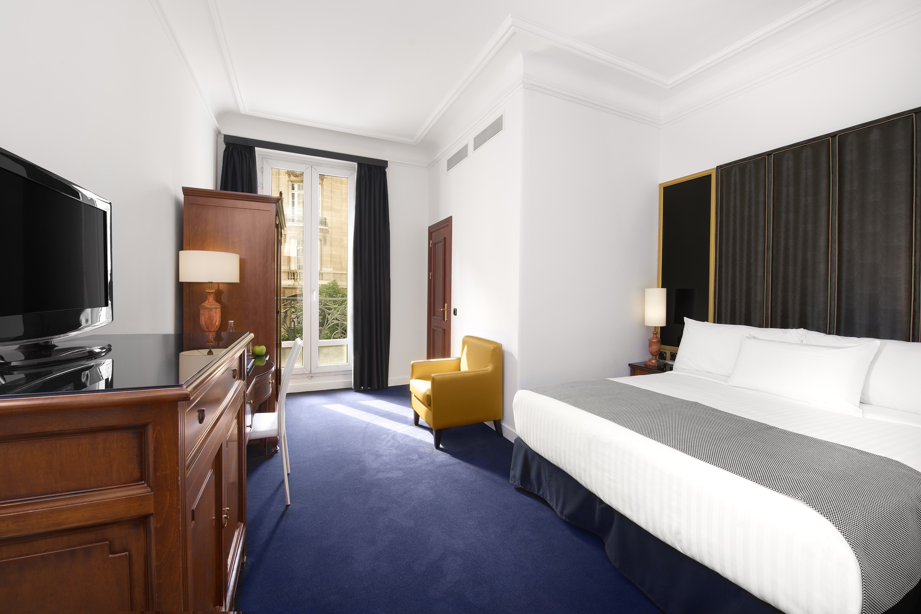 Elegant hotel room with king bed, blue carpet, yellow armchair, and French balcony.