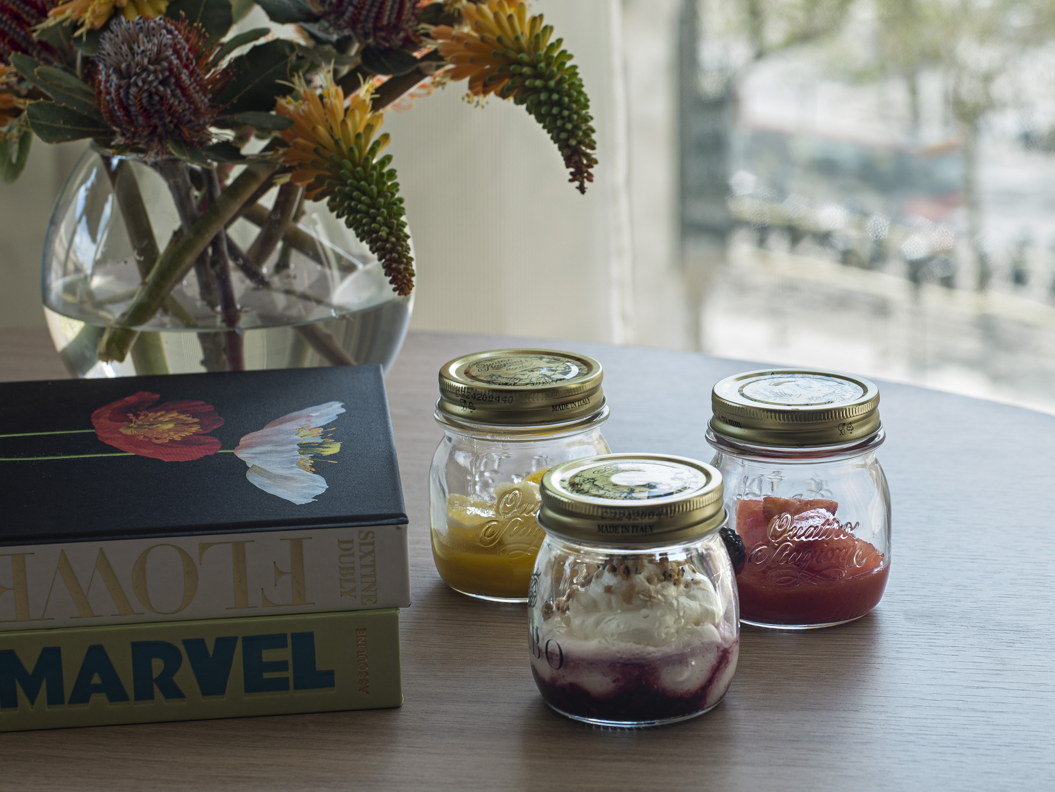 Three layered dessert jars, fruit preserves, flowers, and books on a table.