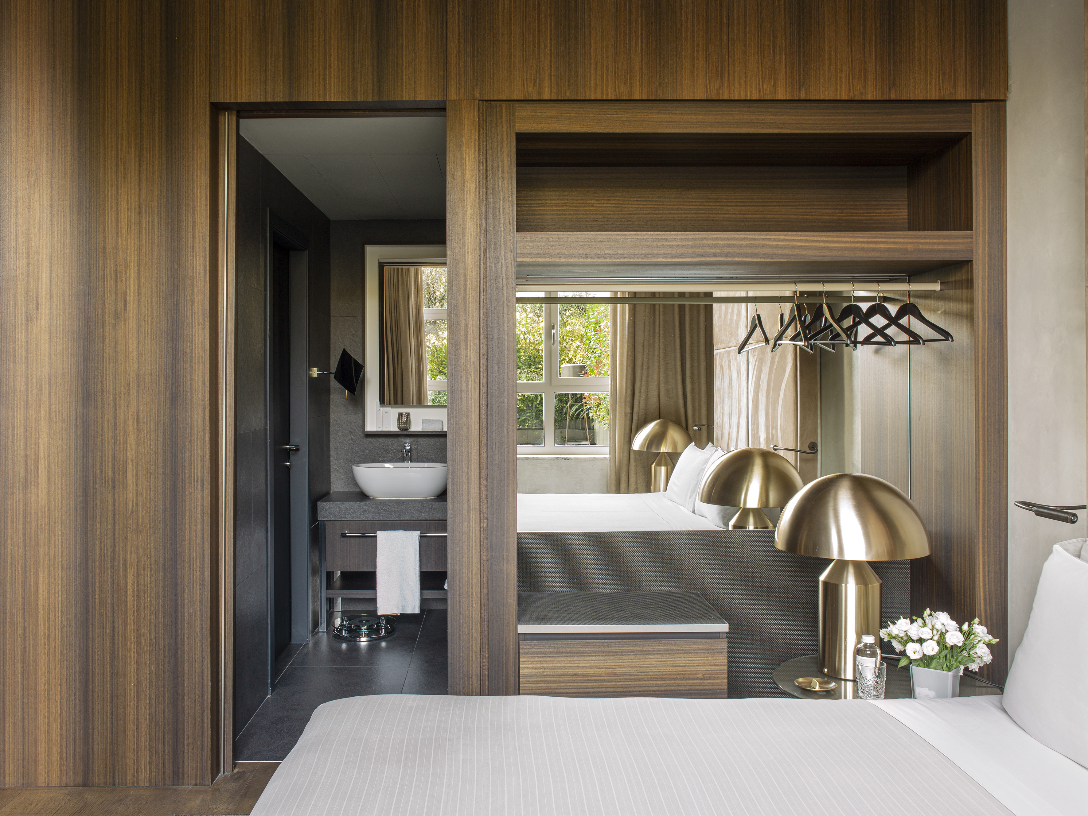 Sleek hotel bedroom with integrated bathroom, wood paneling, and golden bedside lamps.