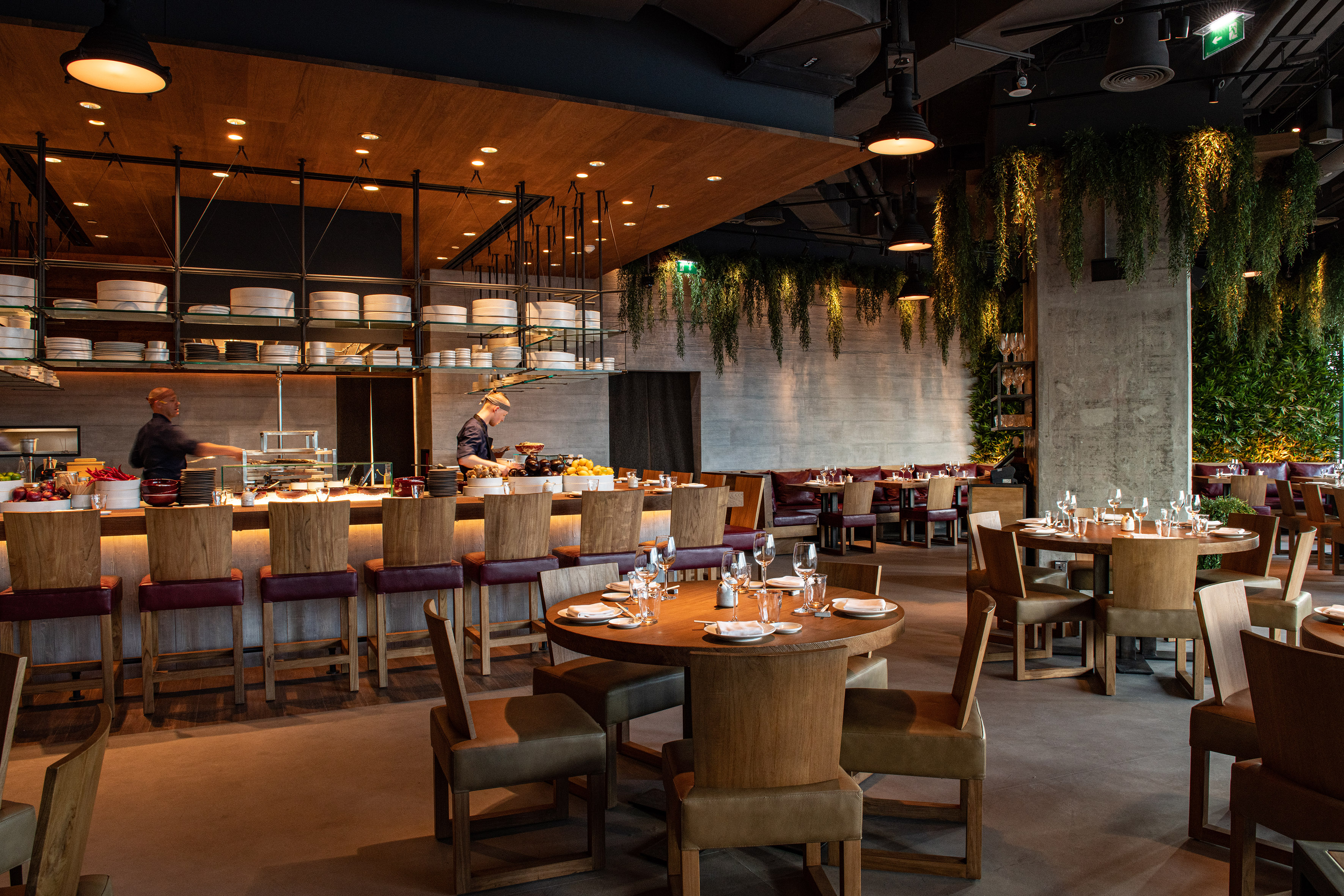 Upscale restaurant with open kitchen, dining tables, and hanging greenery.