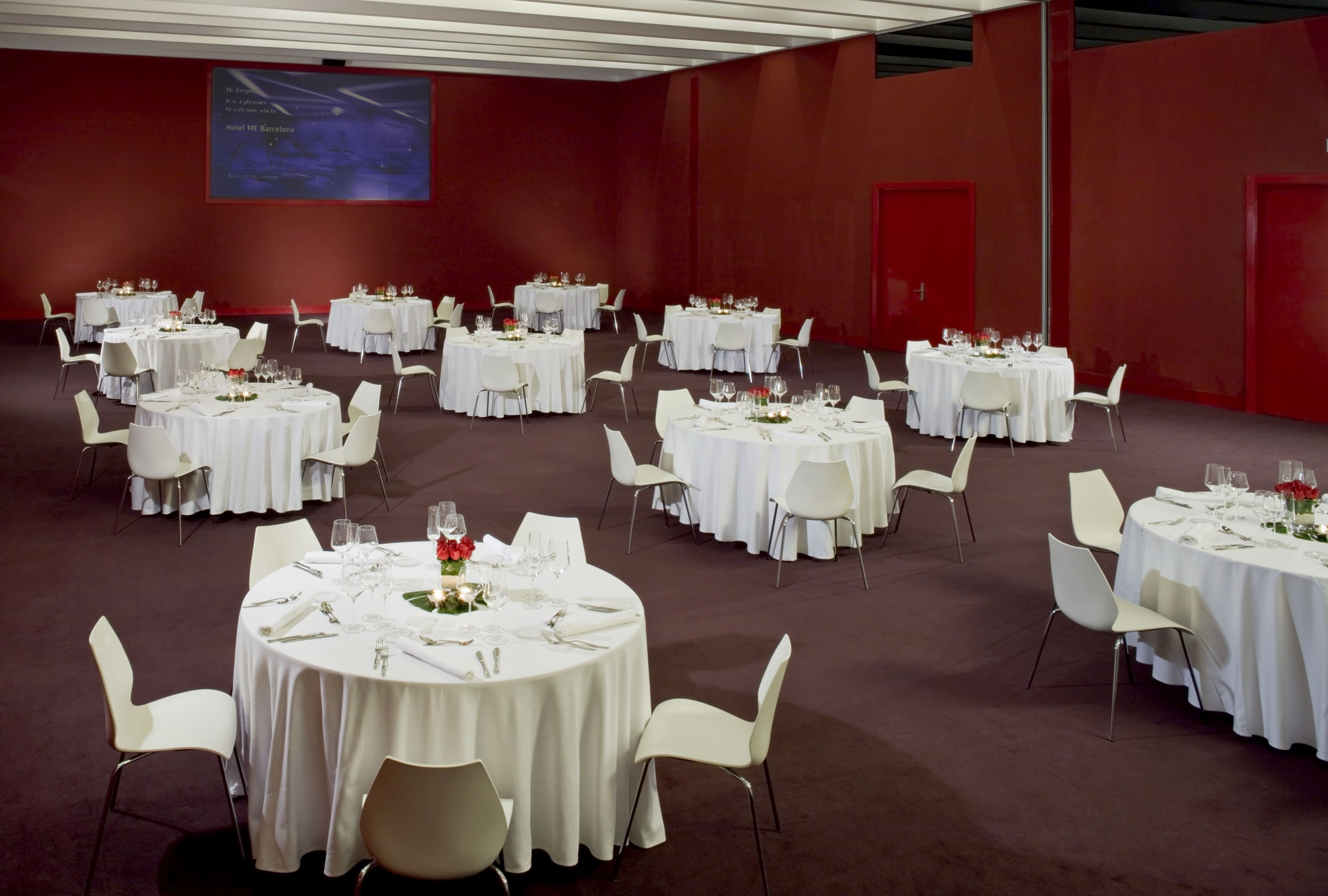 Red hotel event space with white round tables, chairs, and screen display.