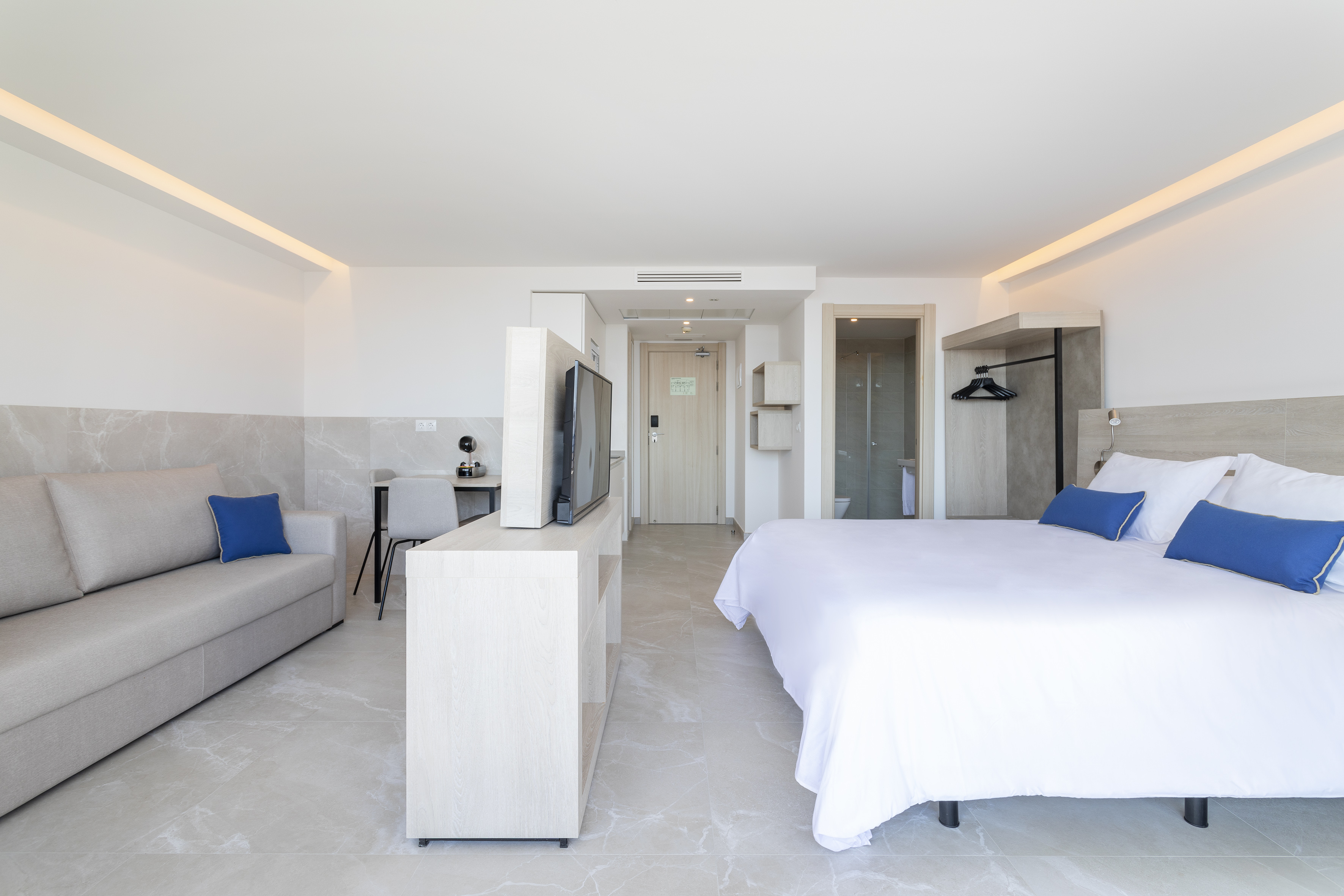 Bright modern hotel room with bed, sofa, desk, and blue accent pillows.
