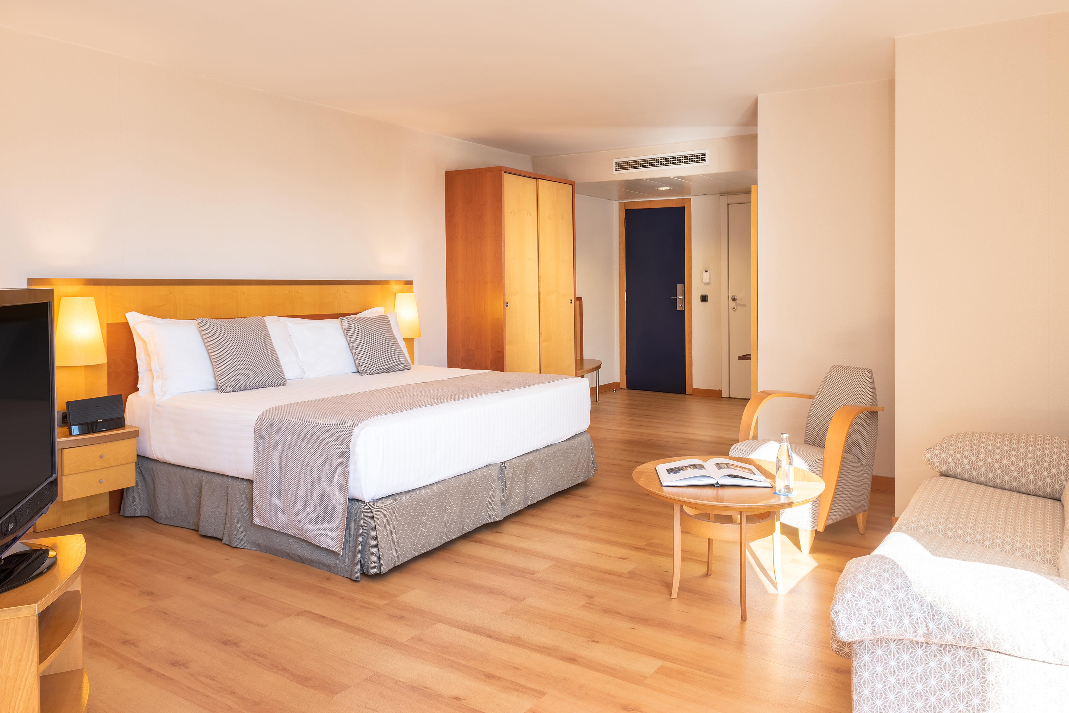 Modern hotel room with king bed, wooden floor, armchair, and sofa.