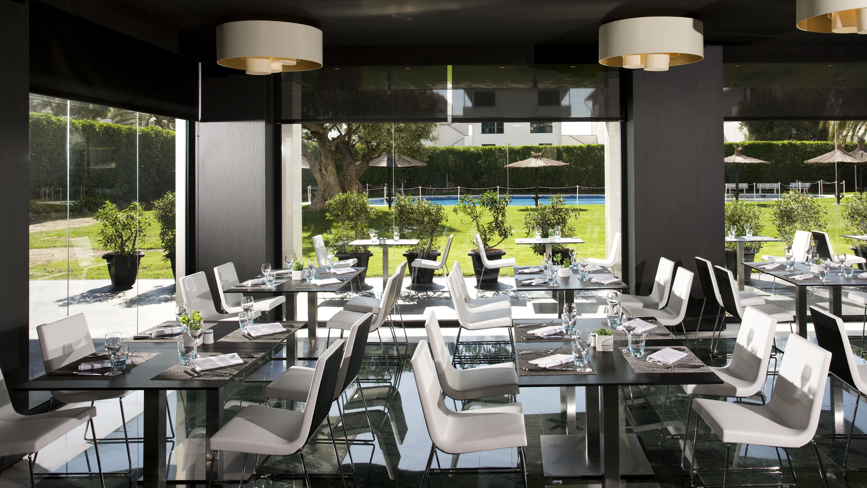 Modern restaurant with black tables, white chairs, and lush garden pool view.
