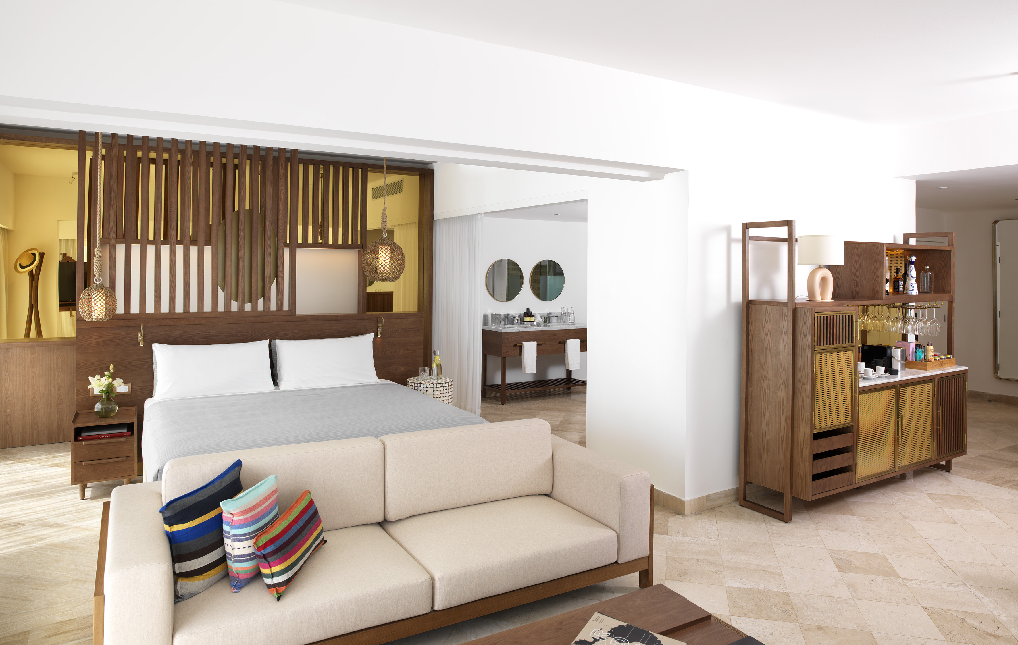 Modern hotel suite with bed, sofa, minibar, and open bathroom area.