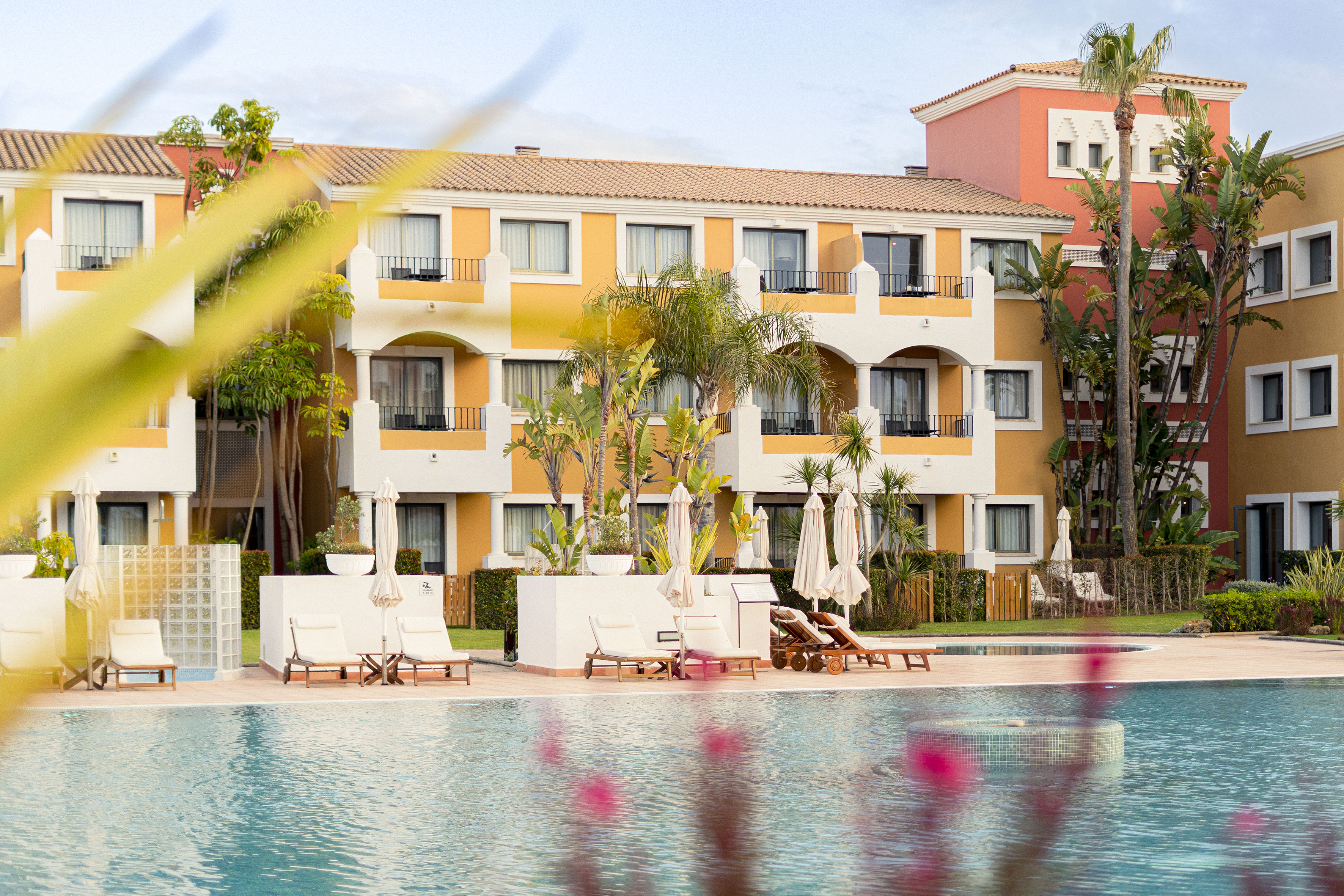 Mediterranean resort hotel with swimming pool, sun loungers, and palm trees.