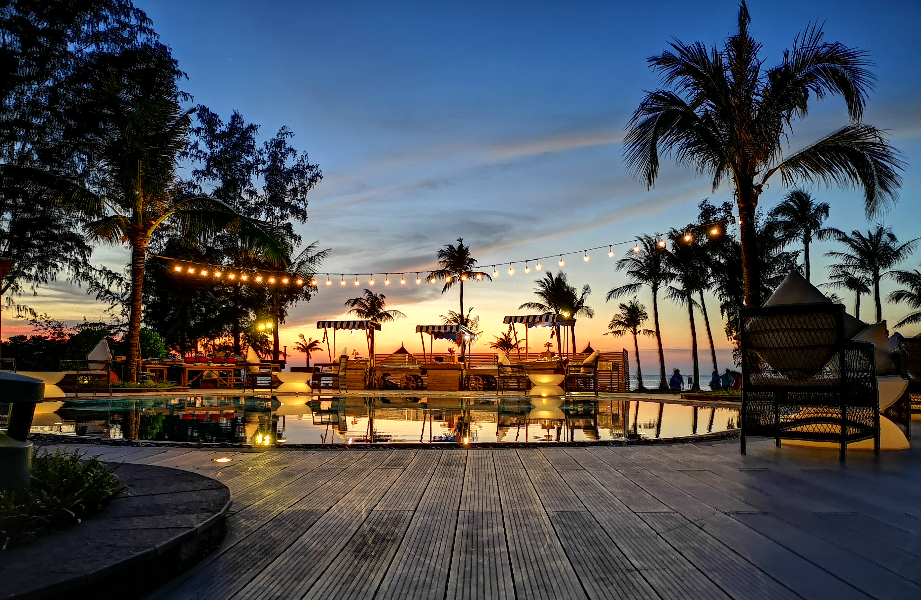 Tropical resort pool deck at sunset with palm trees and string lights.