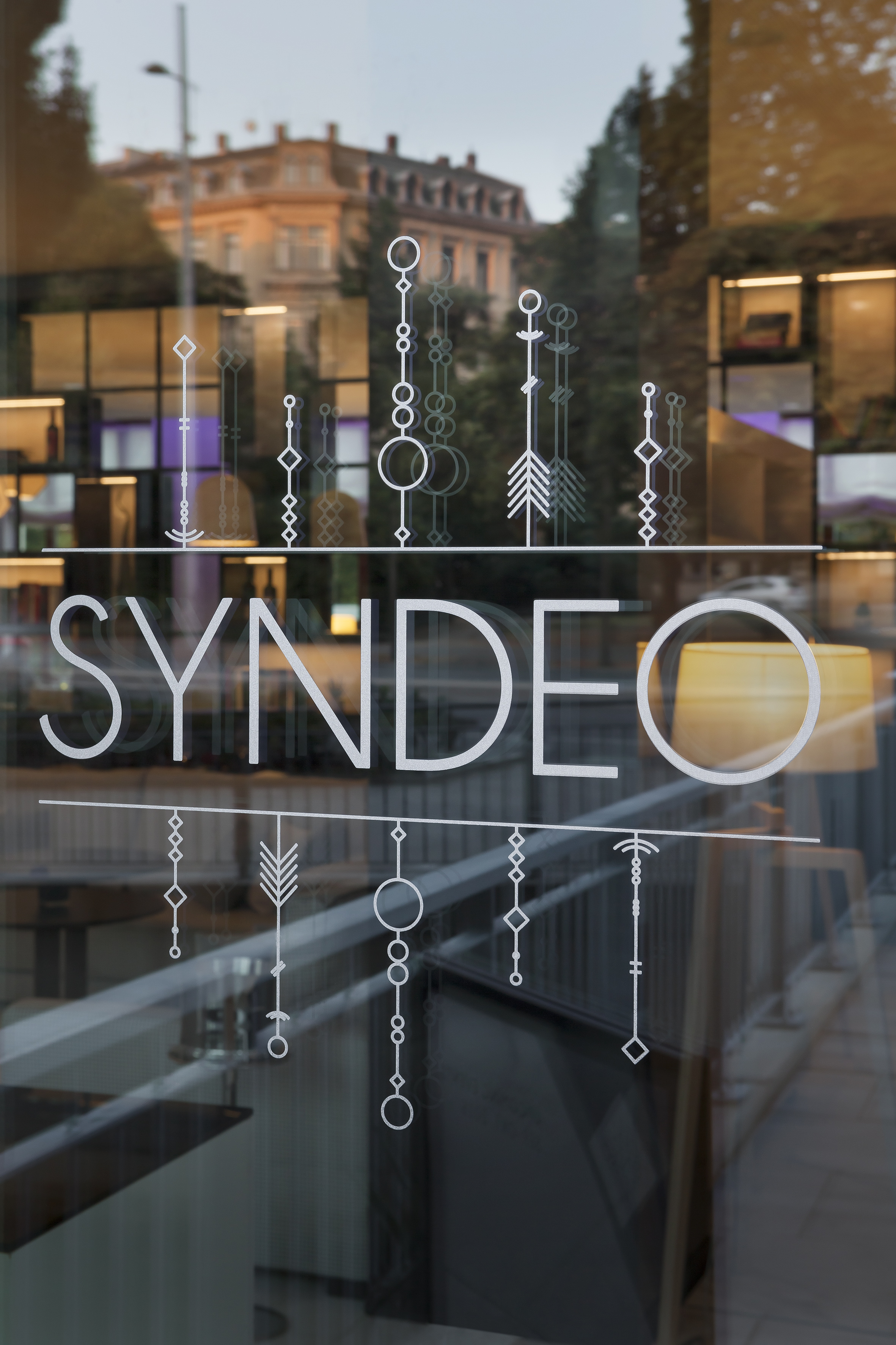 SYNDEO sign on modern glass storefront with geometric patterns, city reflection.