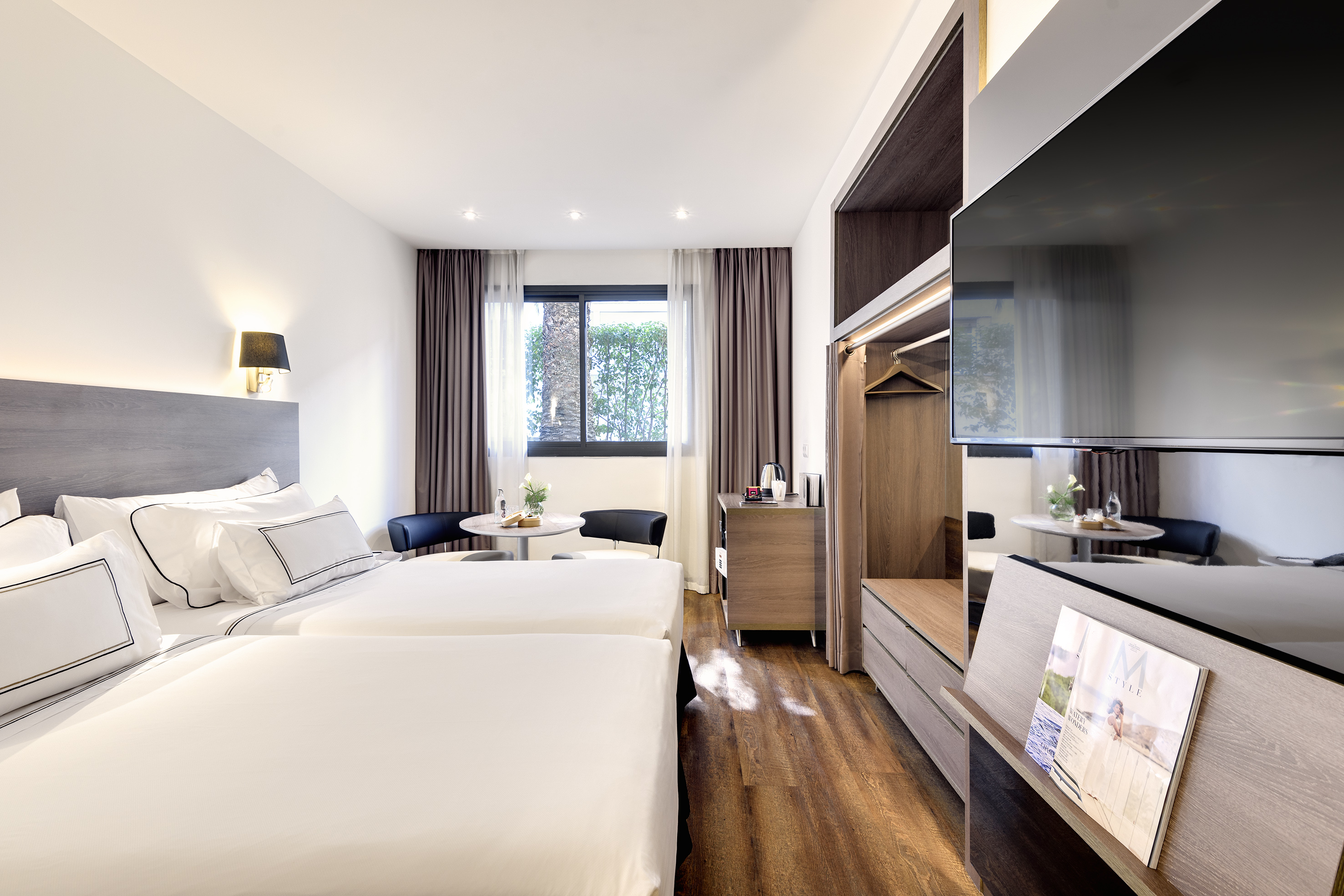 Modern hotel twin room with white beds, window, table, and wall-mounted TV.