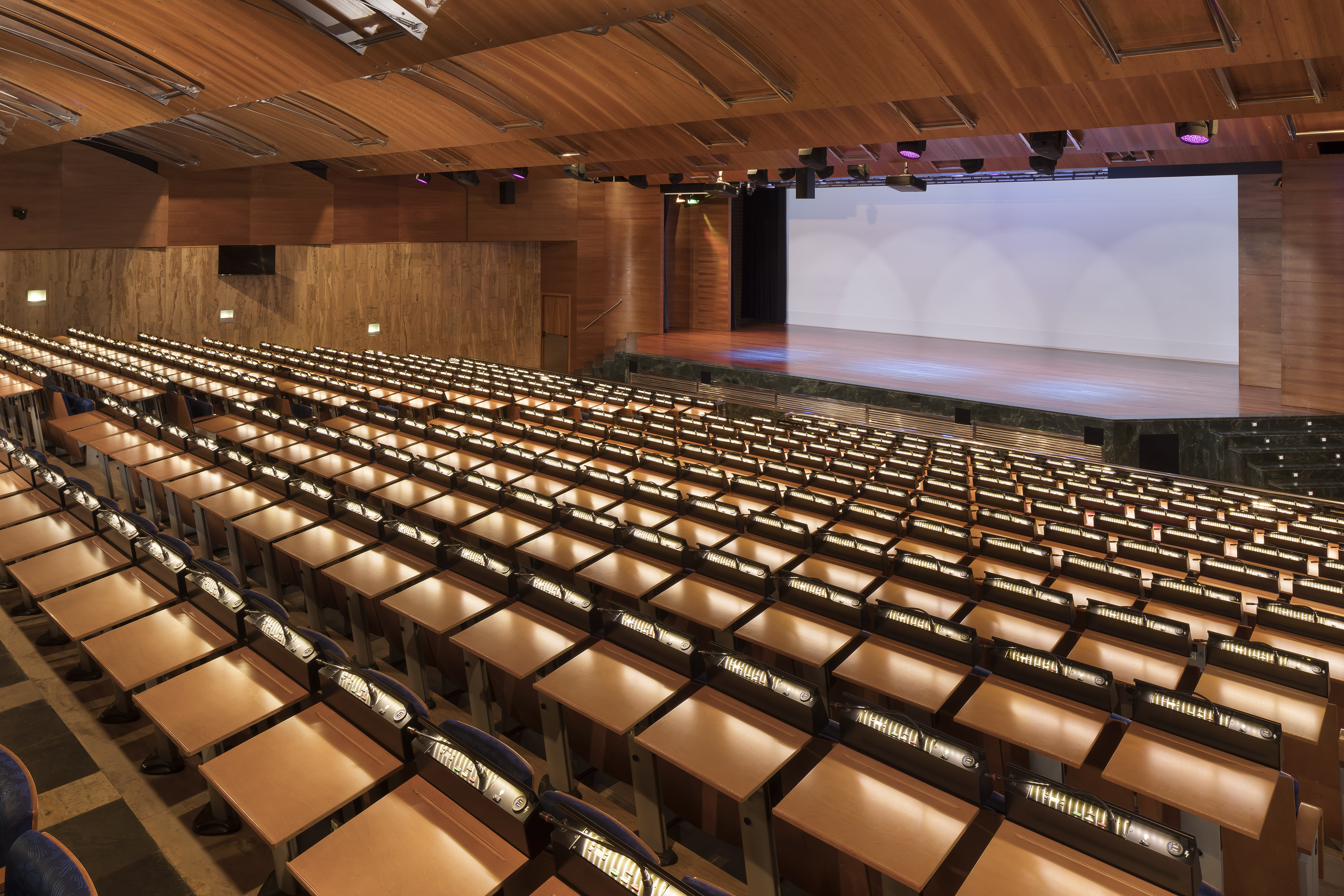 Empty modern lecture hall with tiered wooden desks, stage, and projection screen.