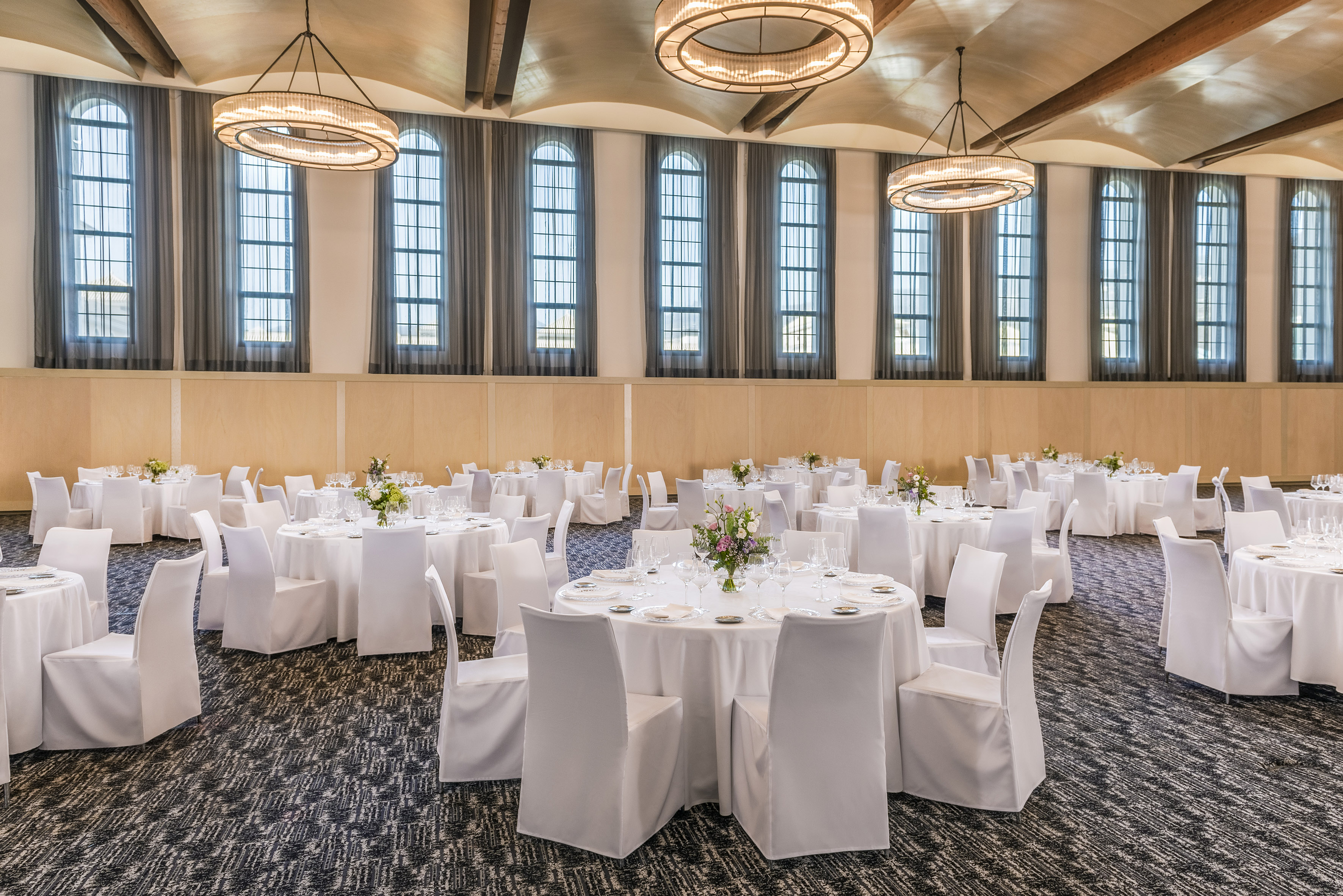 Elegant ballroom with white tables, chairs, arched windows, and chandeliers.