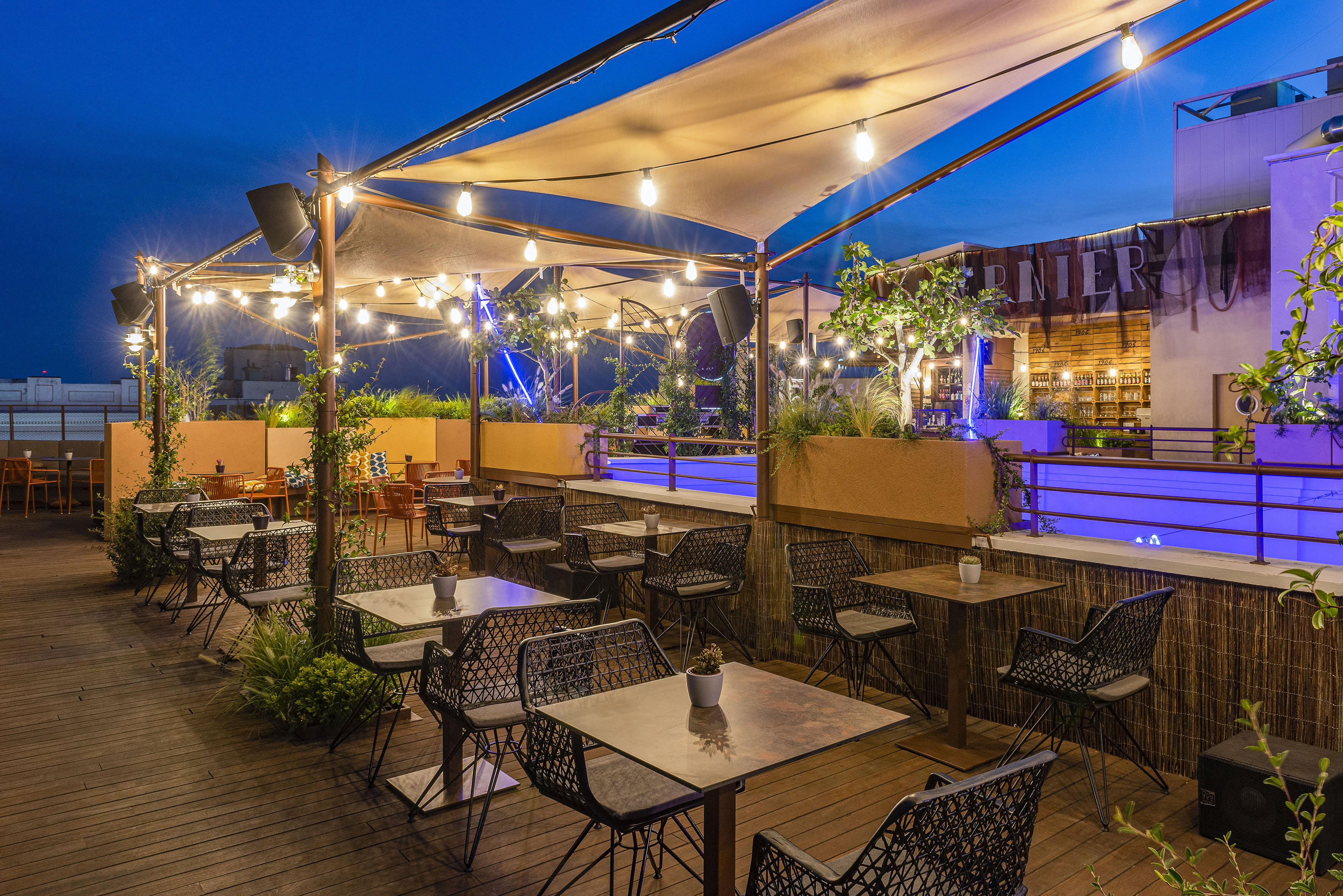 Nighttime rooftop terrace with string lights, outdoor seating, and blue bar.
