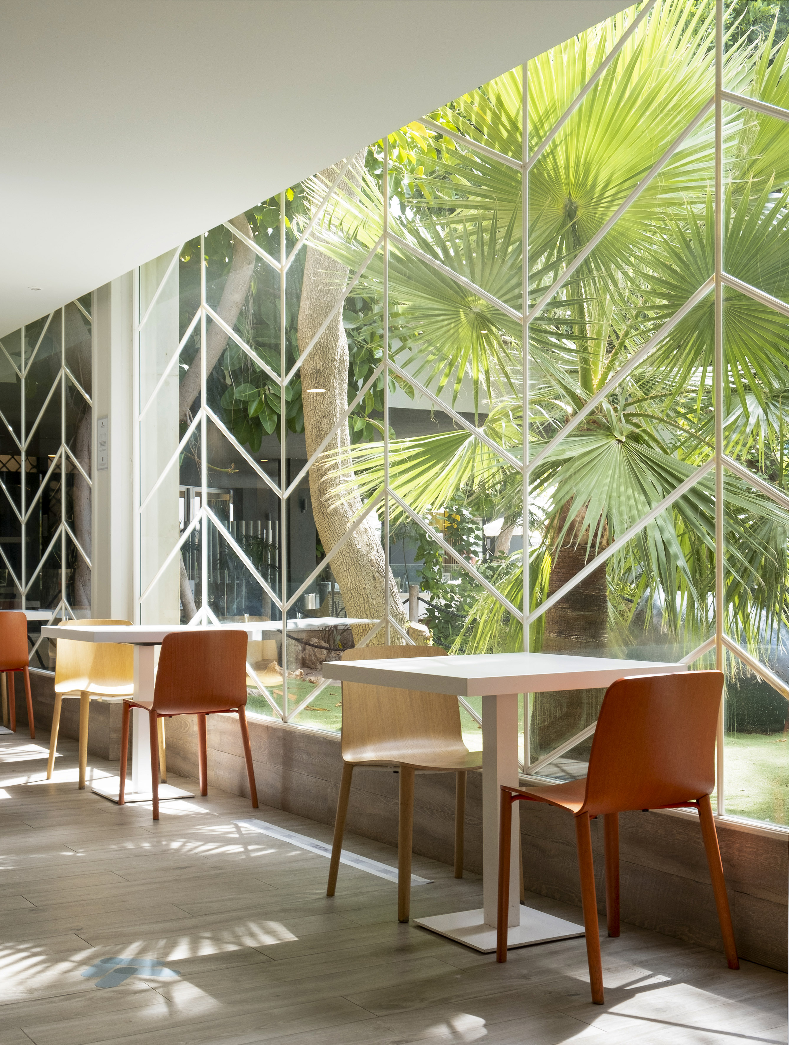 Modern sunlit cafe with geometric window, white tables, and views of palm trees.