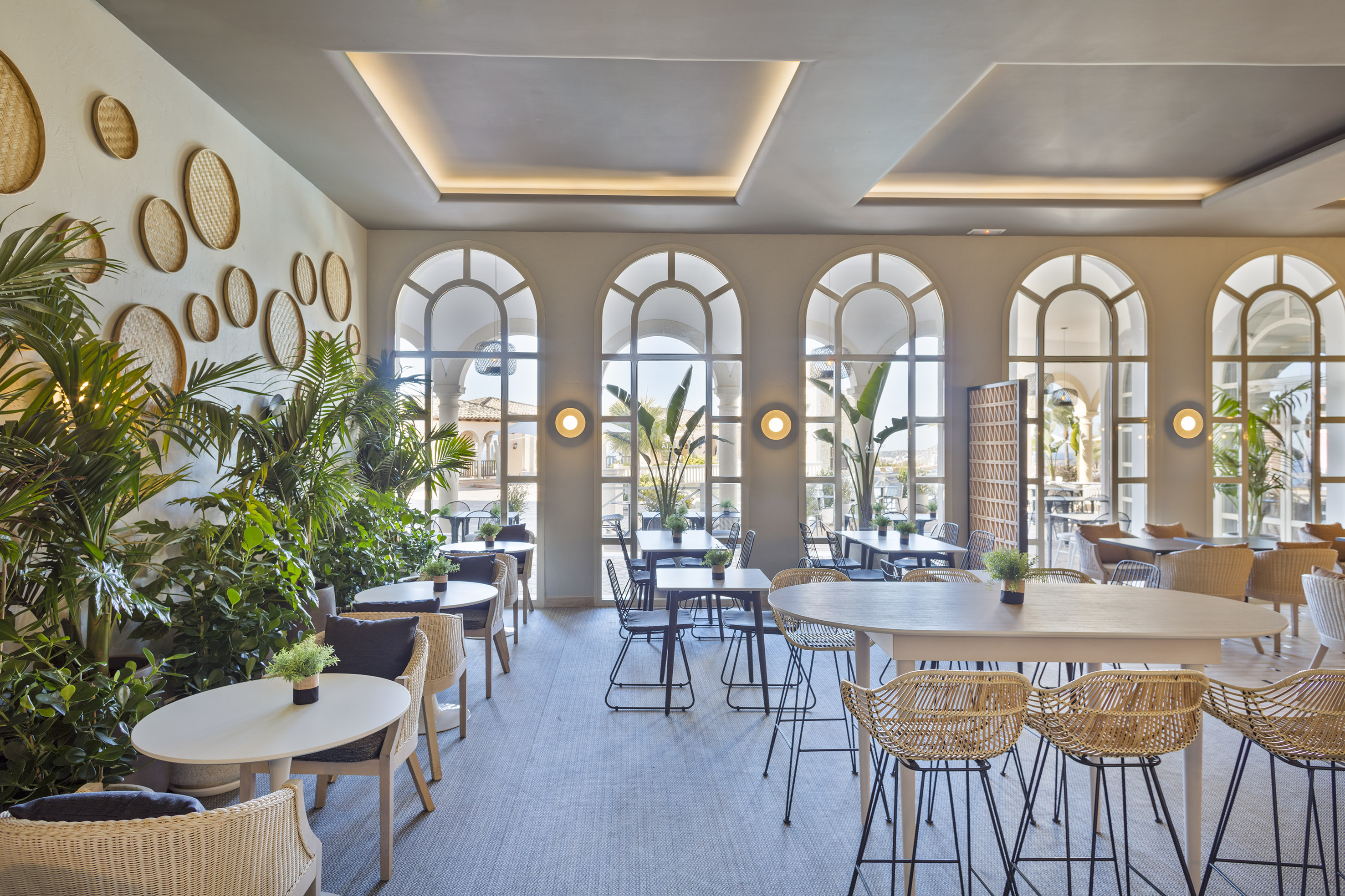 Bright restaurant interior with arched windows, lush plants, and wicker chairs.