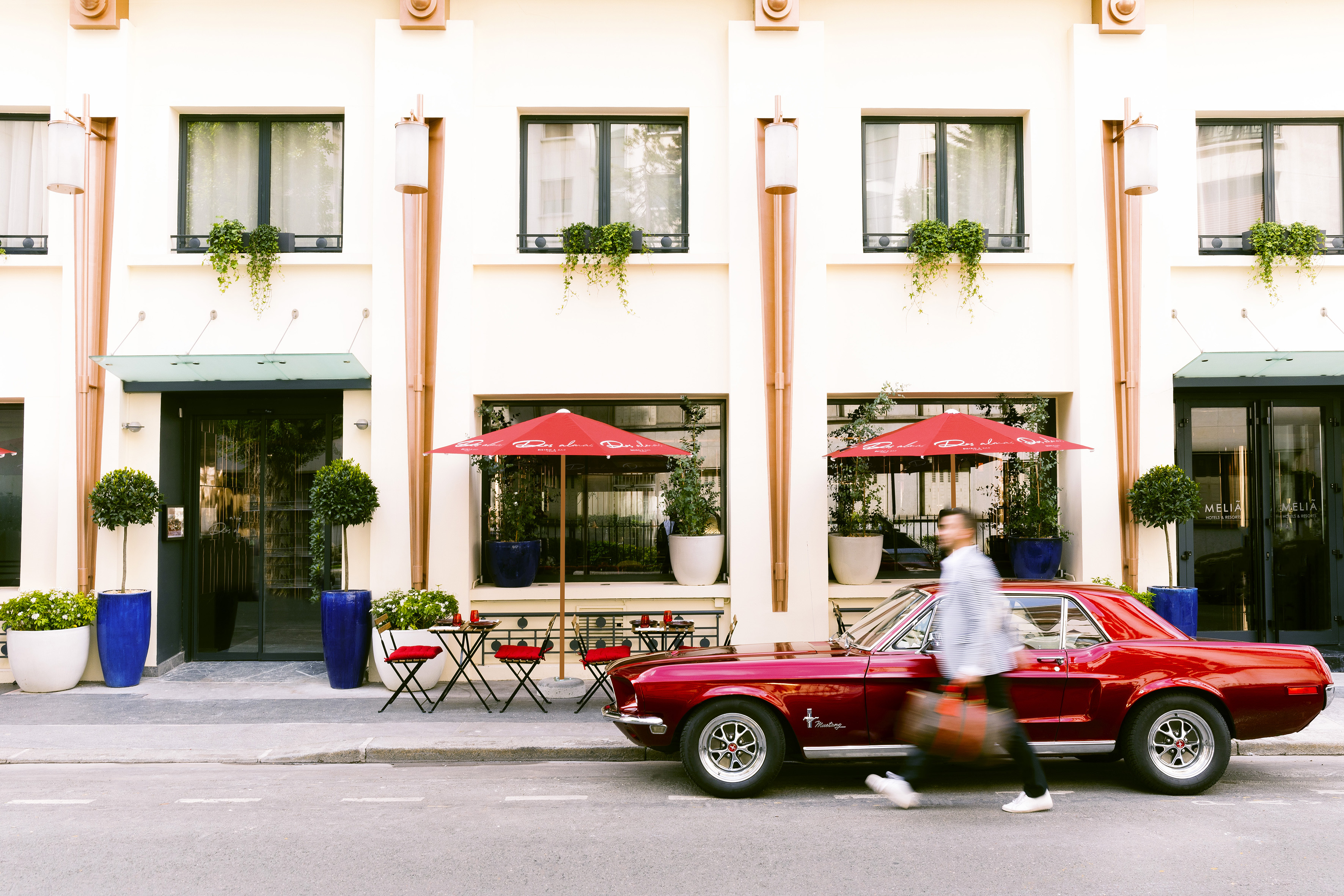 Man walking past red classic Mustang, hotel exterior with outdoor cafe.