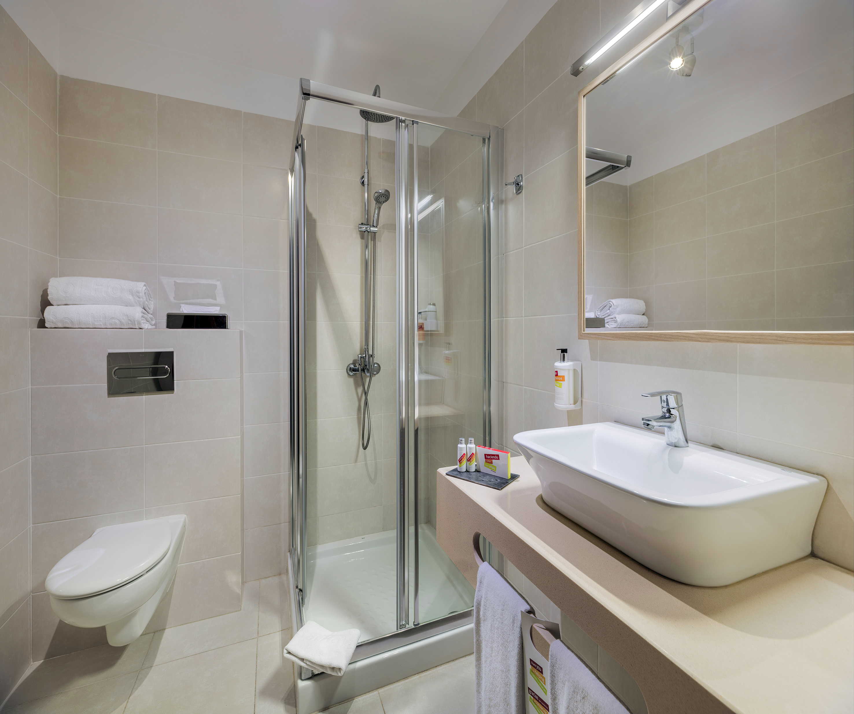 Modern hotel bathroom with shower, toilet, sink, mirror, and beige tiles.