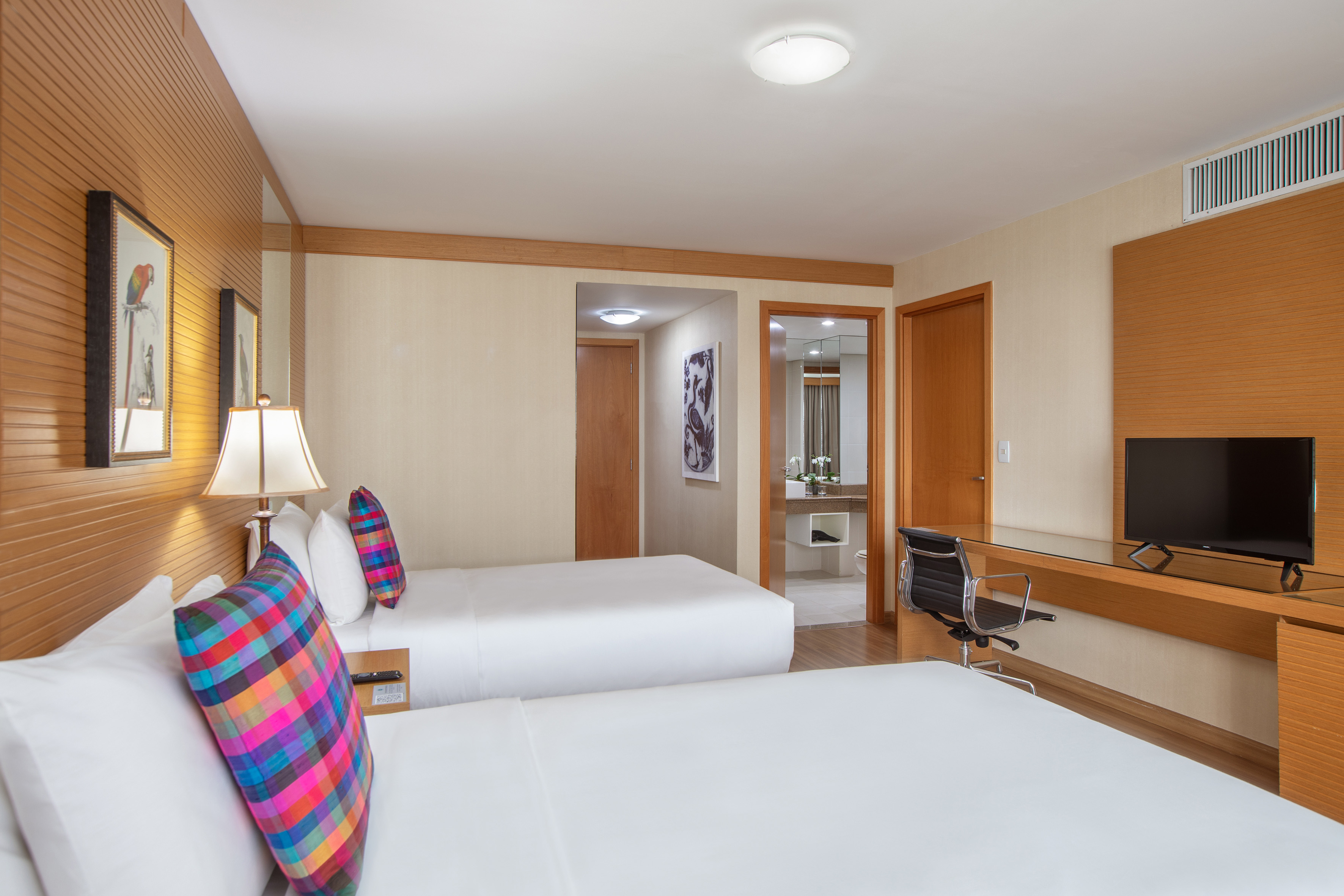Hotel room with two beds, colorful pillows, desk, TV, and bathroom entrance.