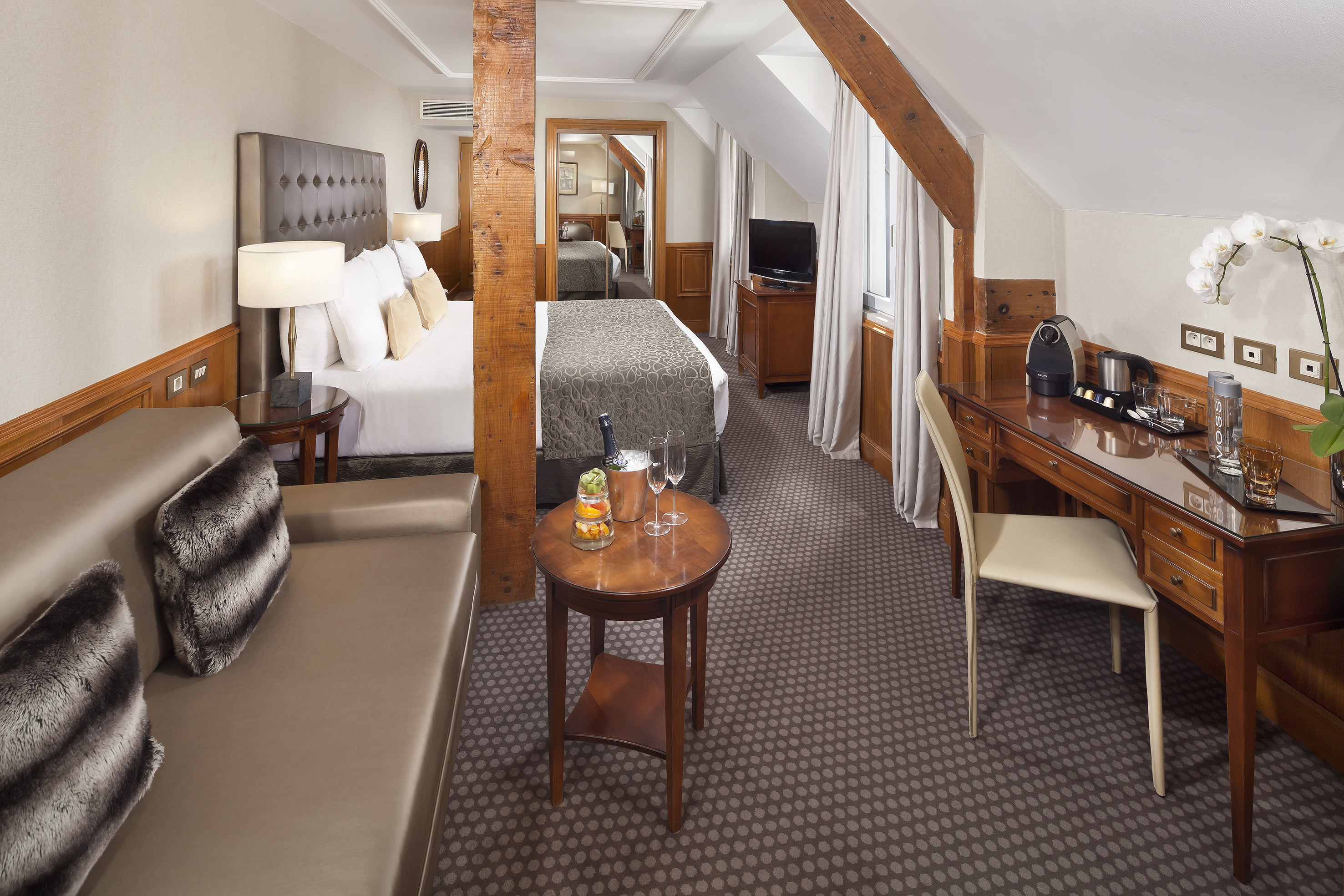 Elegant hotel room with bed, sofa, desk, champagne, coffee, and exposed beams.