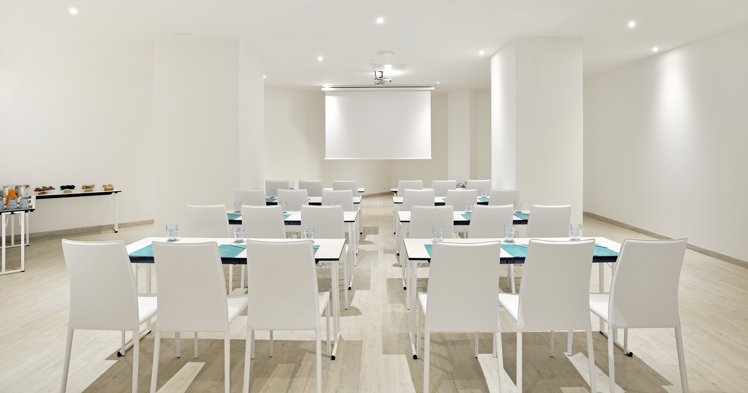 Modern event space with white tables, chairs, projector screen, and refreshments.