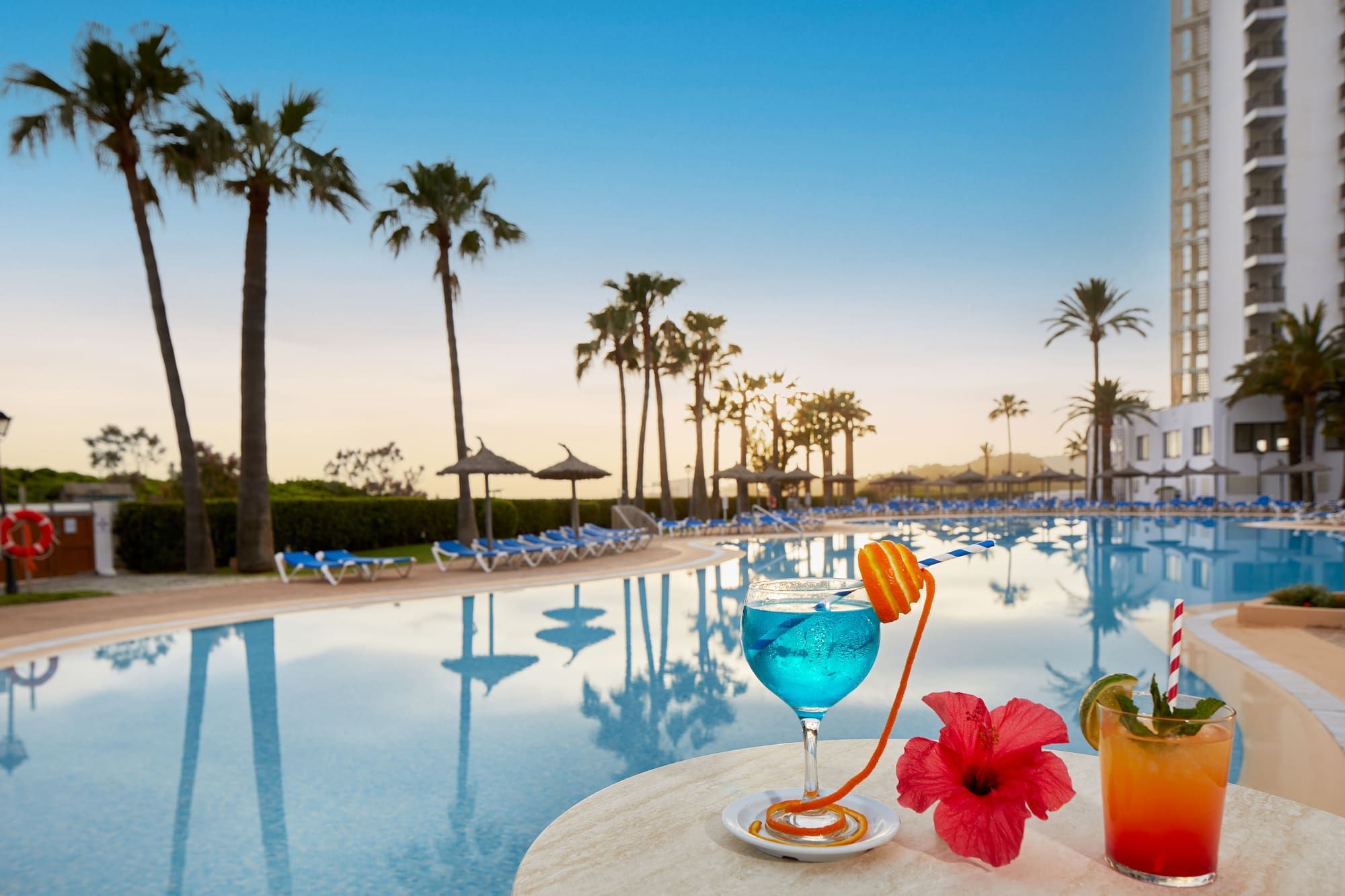 Tropical cocktails by a hotel swimming pool with palm trees at sunset.