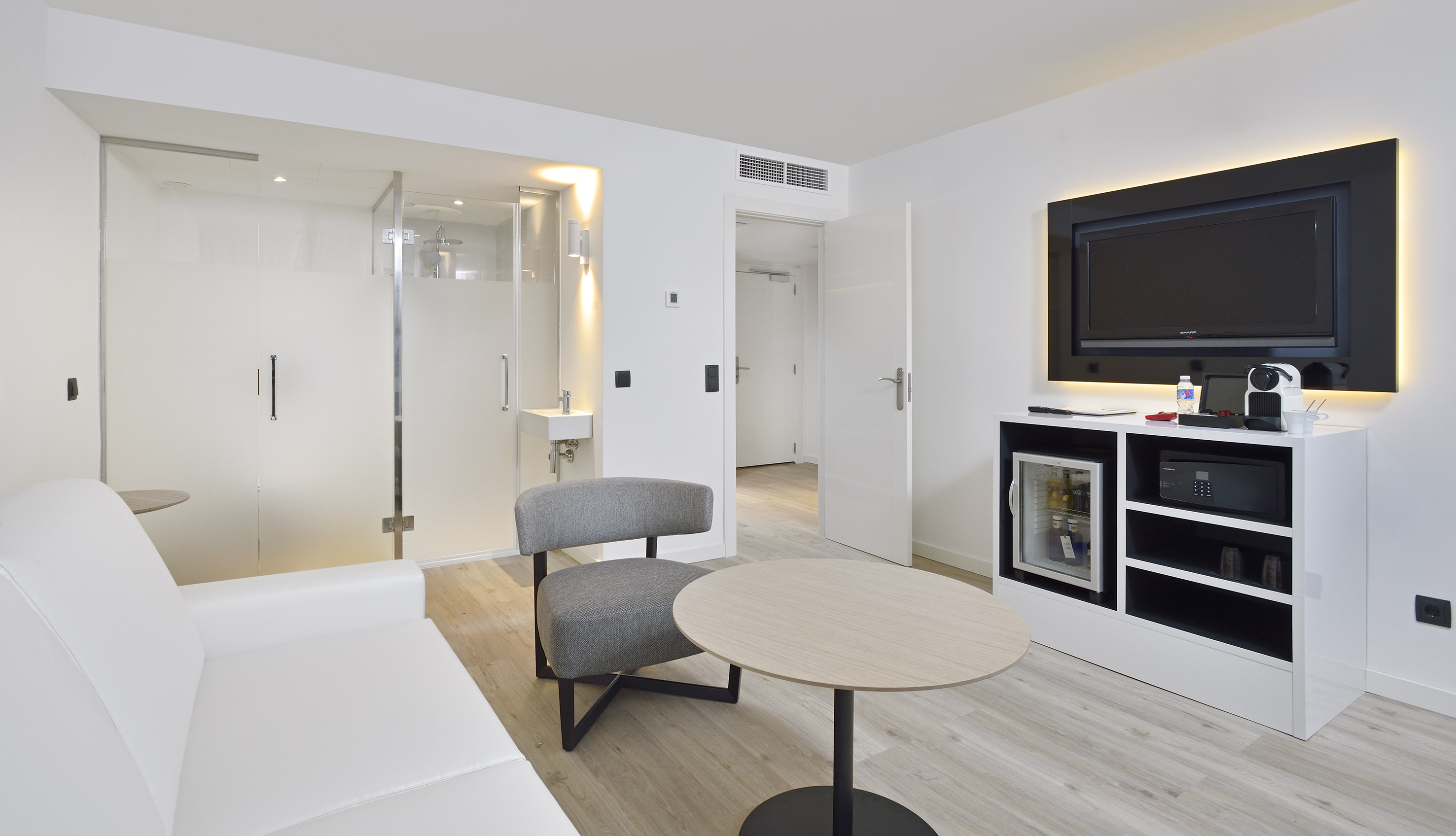 Modern hotel room suite with white decor, TV, mini-fridge, and glass shower.