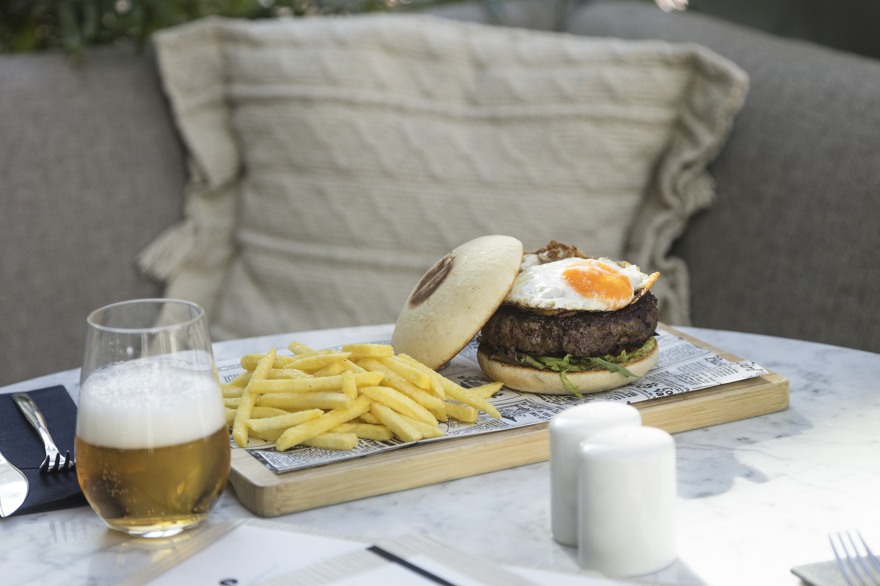 Burger with fried egg, French fries, and beer on a marble table.