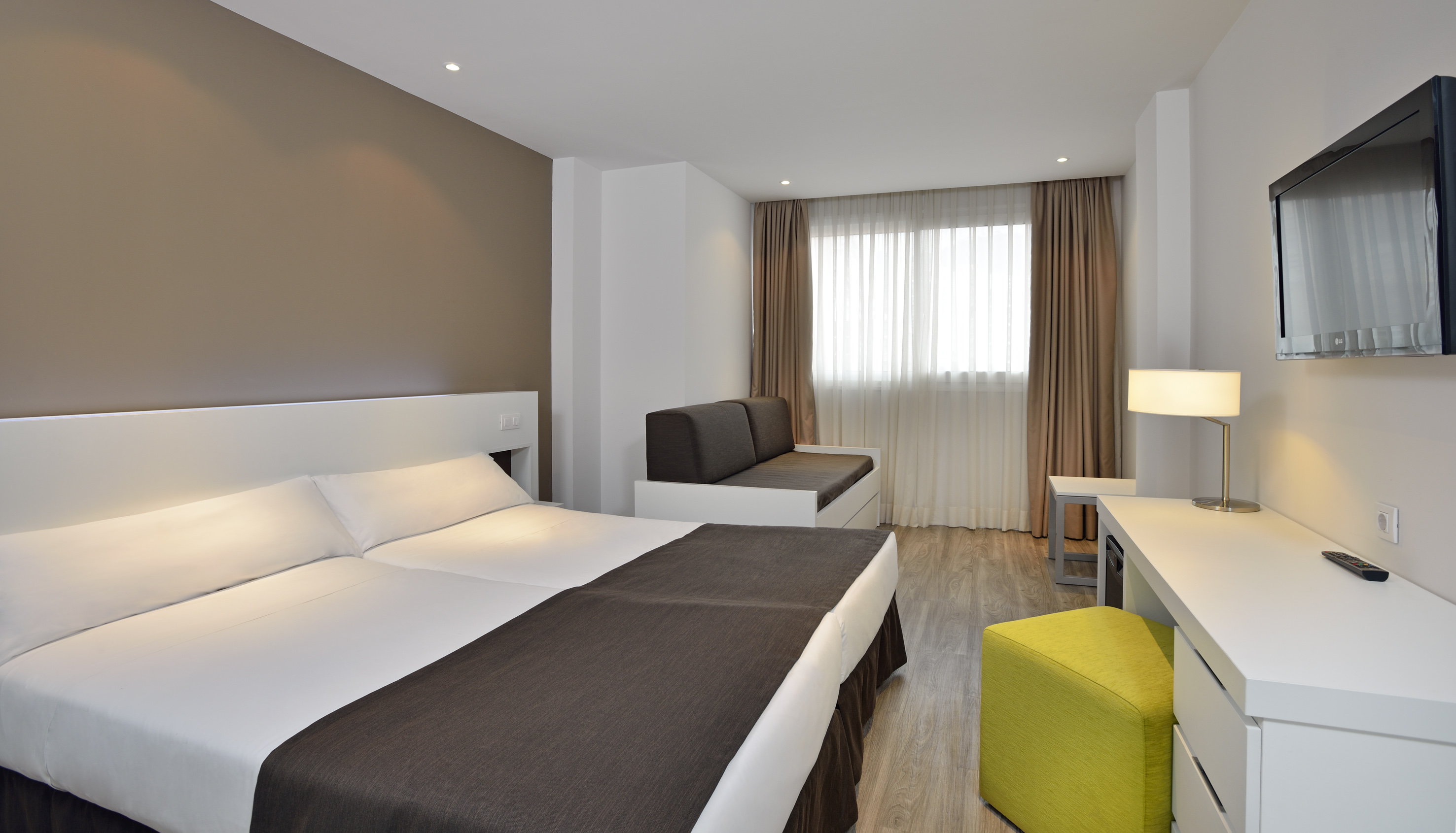 Modern hotel room with twin beds, sofa, white desk, and wall-mounted TV.