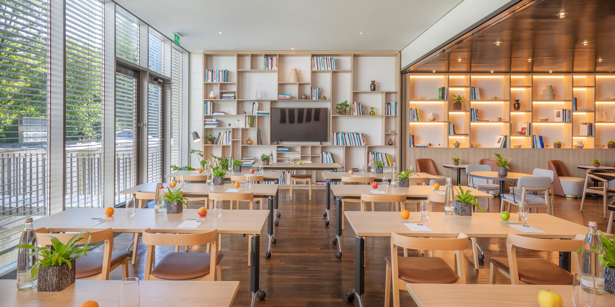 Modern training room with wood tables, natural light, bookshelves, and TV.