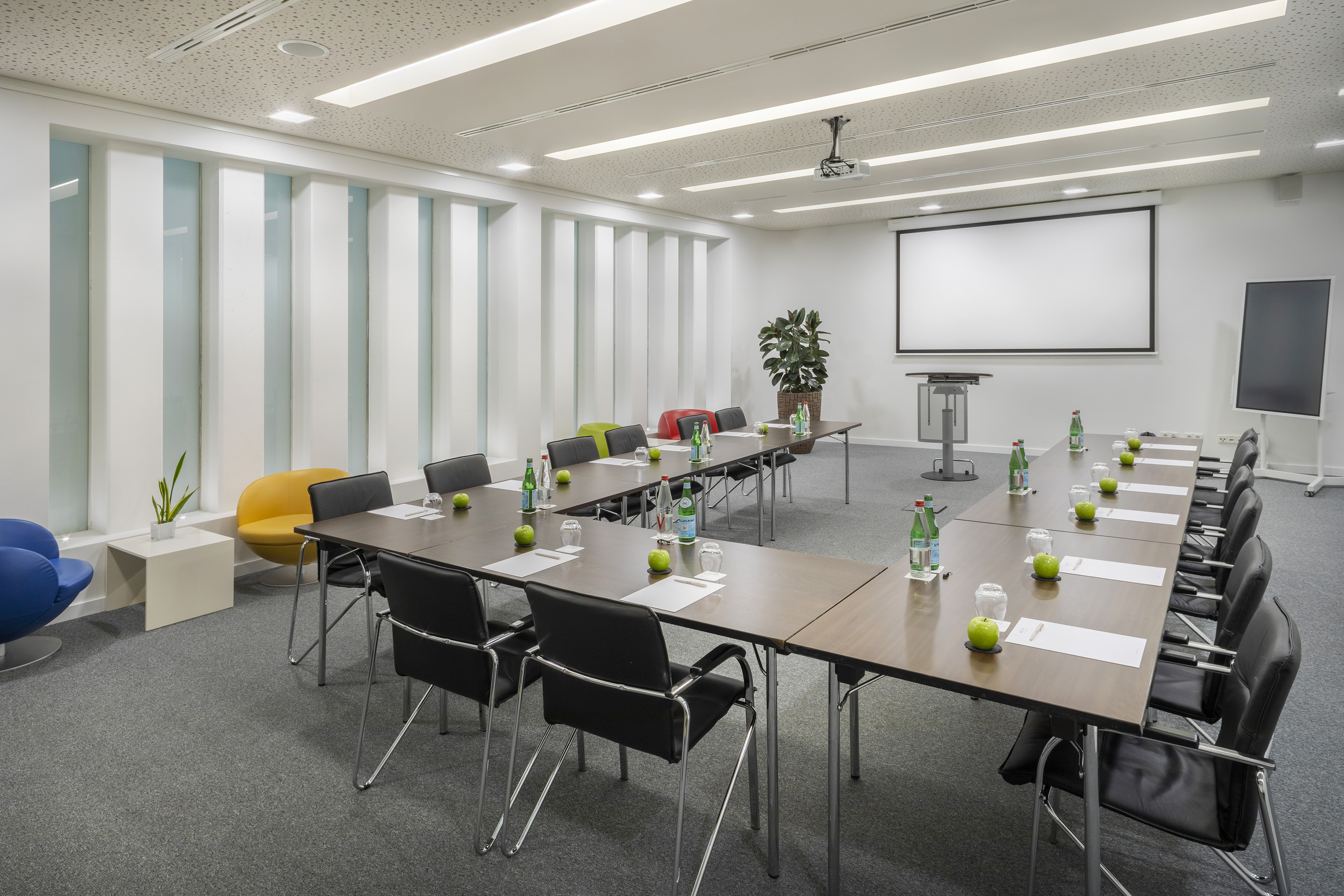 Modern meeting room with U-shaped table, projector screen, water and apples.