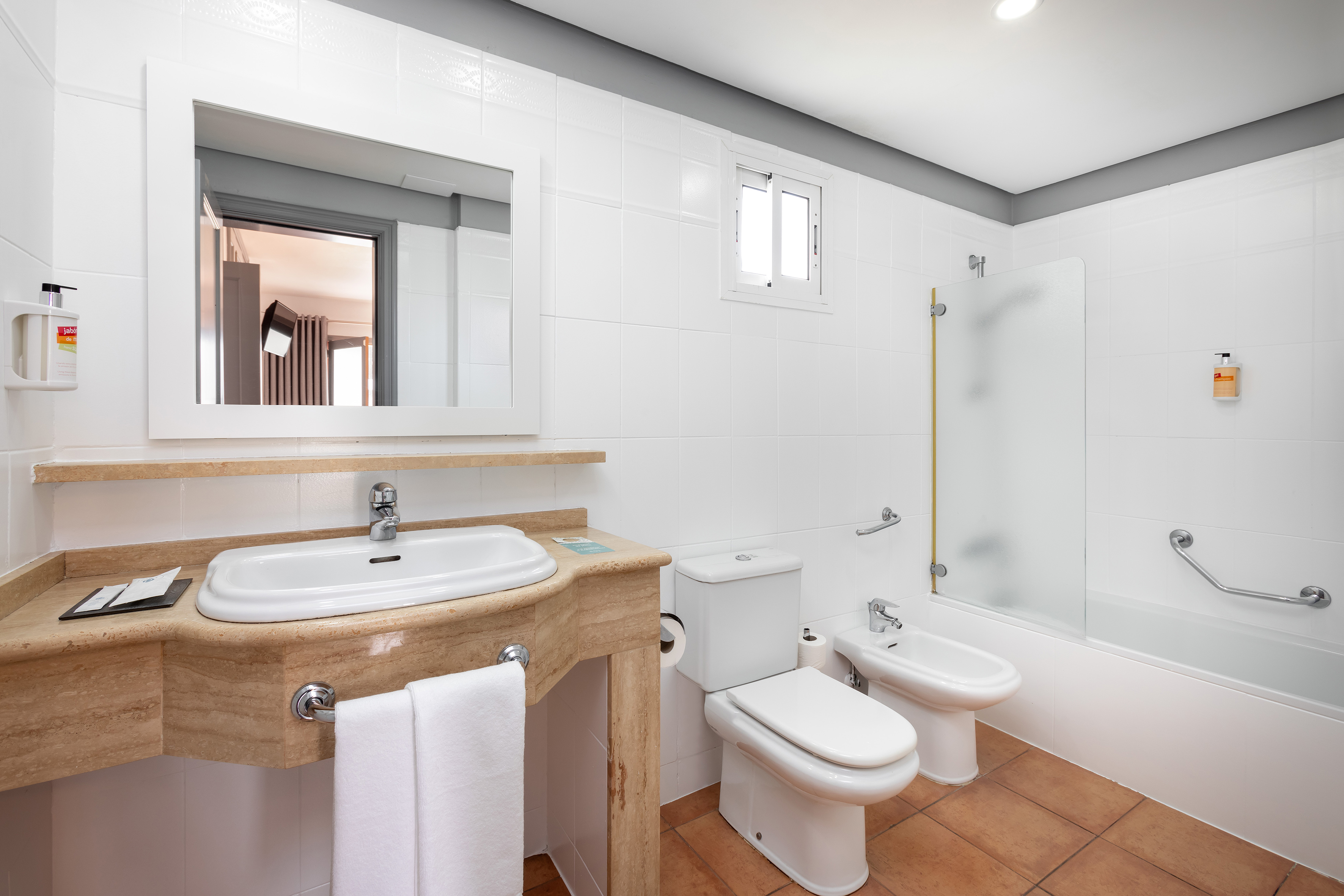 Modern hotel bathroom with white tiles, sink, toilet, bidet, and tub.